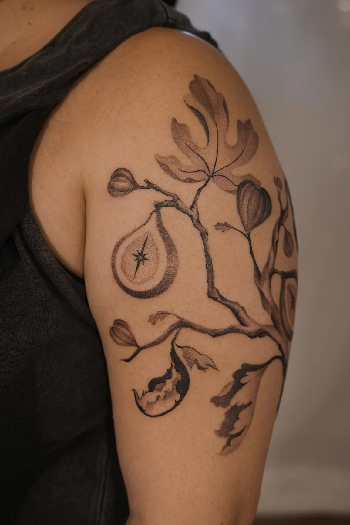 A person with a black and gray branch tattooed on their upper arm, featuring leaves, buds, a teardrop shape with a star inside, and a crescent moon with a face.