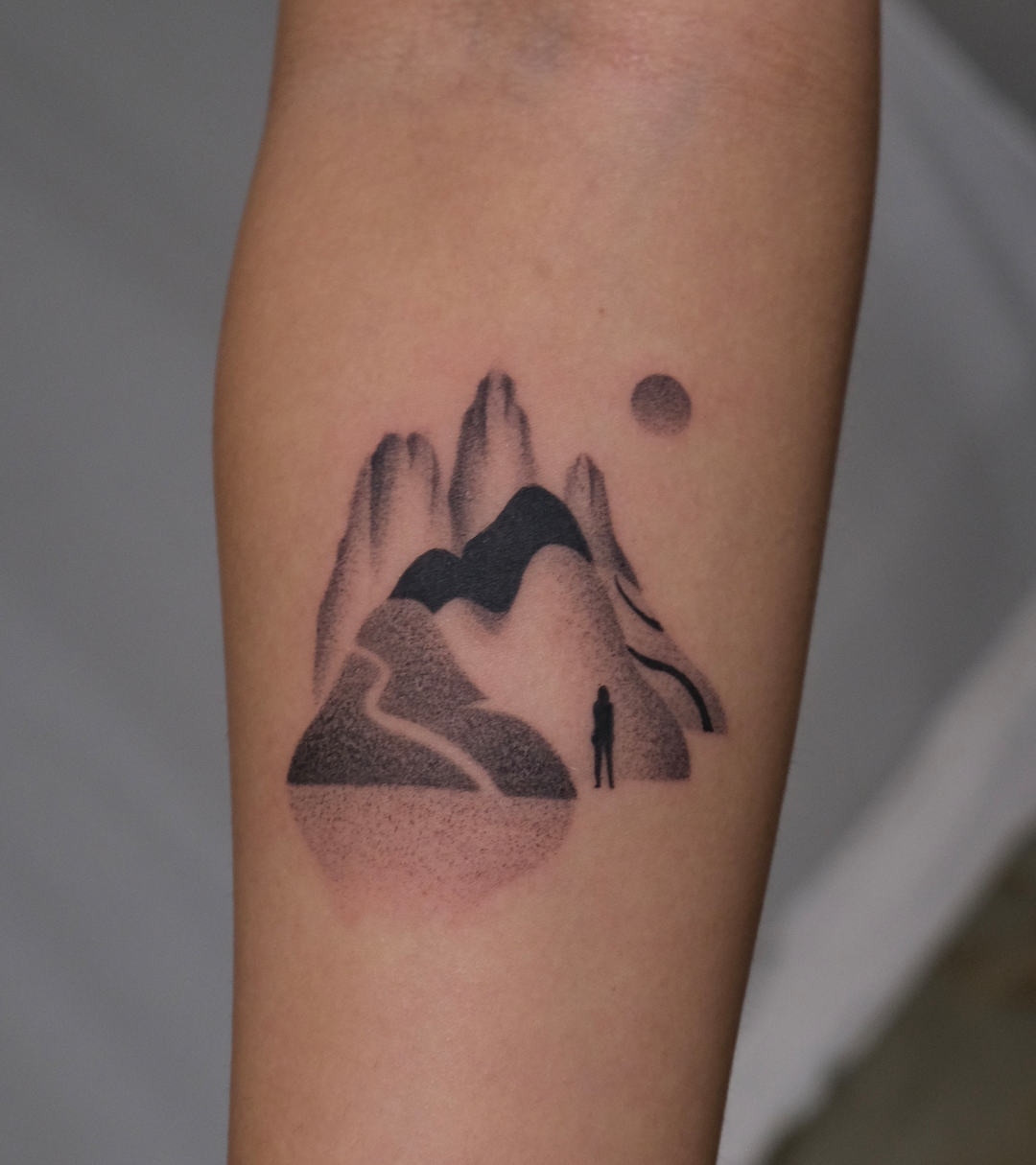 Tattoo of mountains, a person standing, and a sun in black ink on a person's arm, contemporary and unique by New York based contemporary tattoo artist, Ginseng Ink.