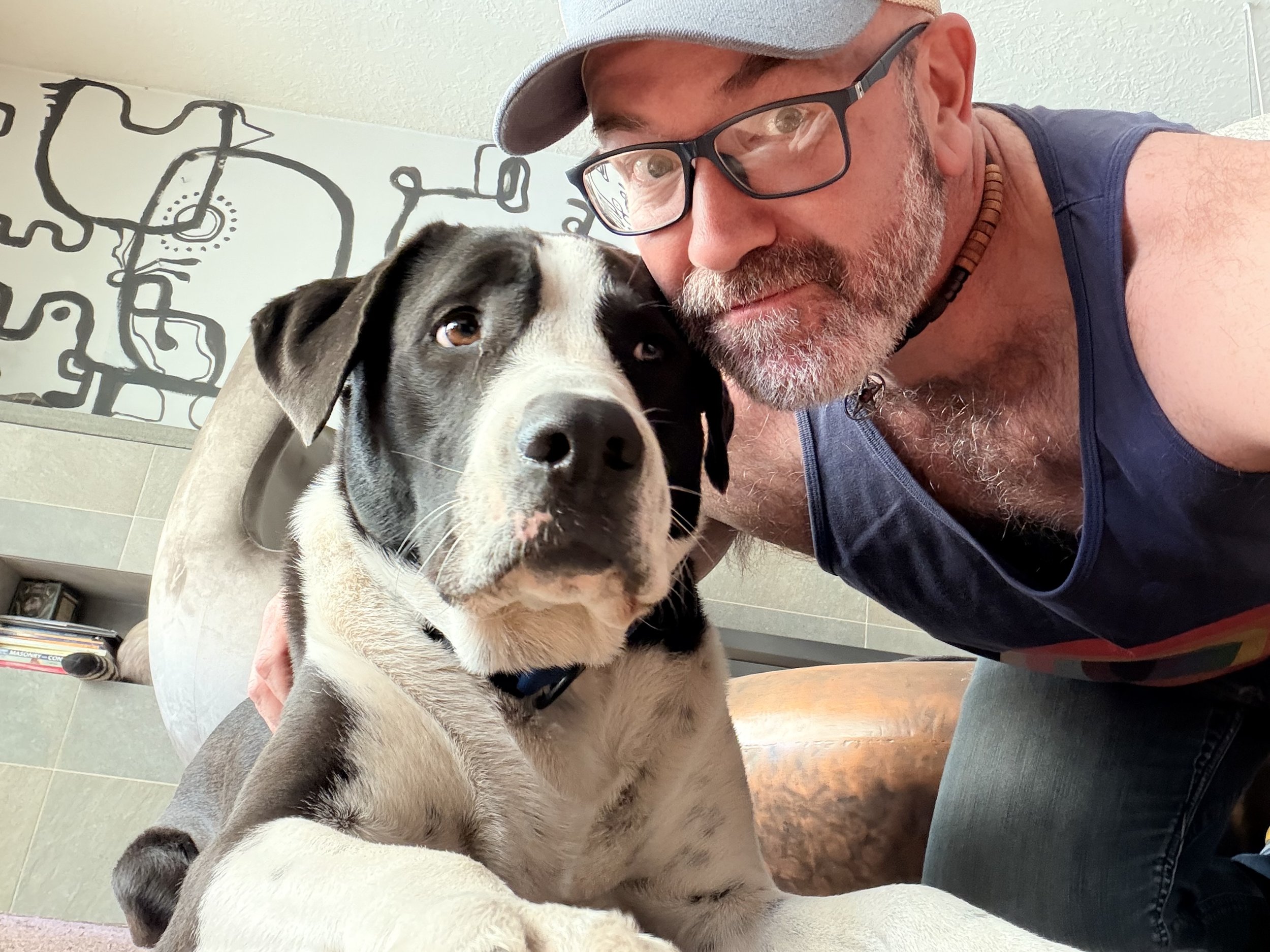 A man with glasses and a gray cap taking a selfie with a black and white puppy on his lap inside a room.