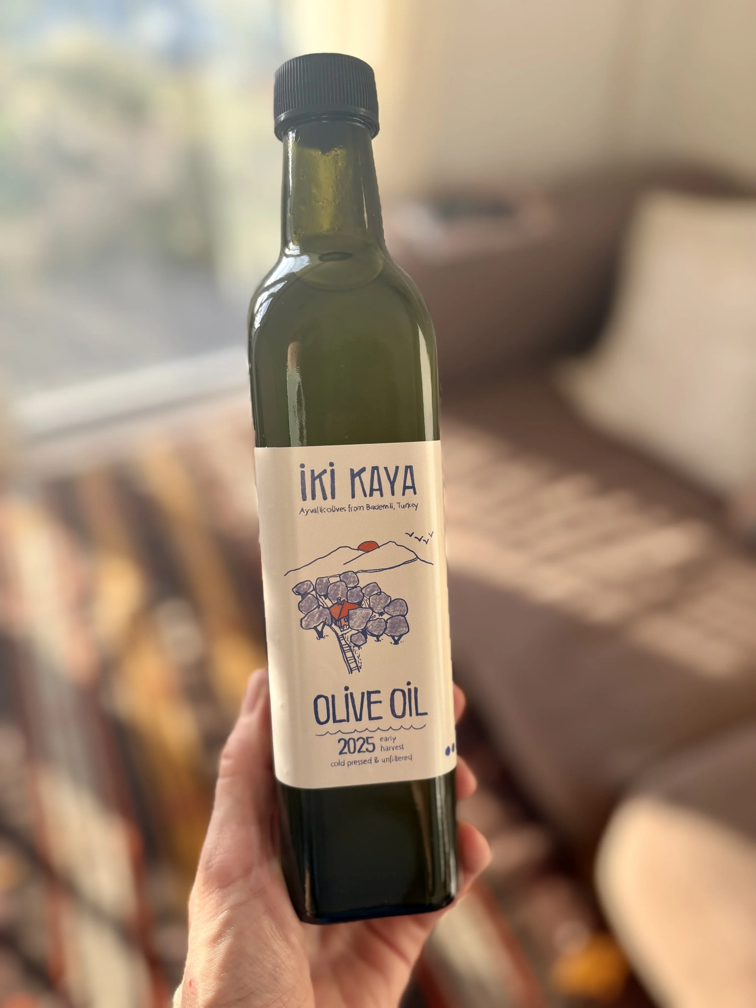 A person holding a dark green bottle of Iki Kaya olive oil with a white label featuring a mountain sketch and text, in a blurry indoor setting.
