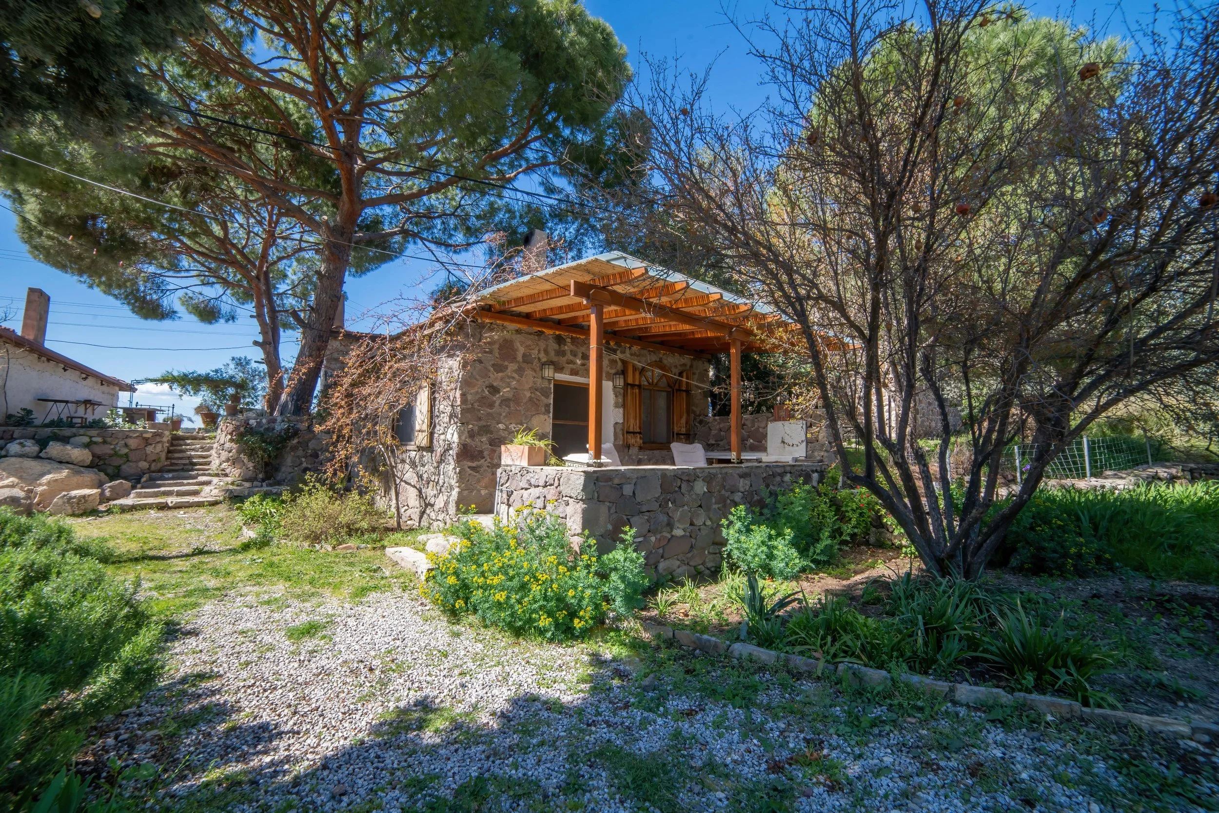 A small stone house with a wooden porch surrounded by trees and greenery in a garden with a gravel path and plant beds.