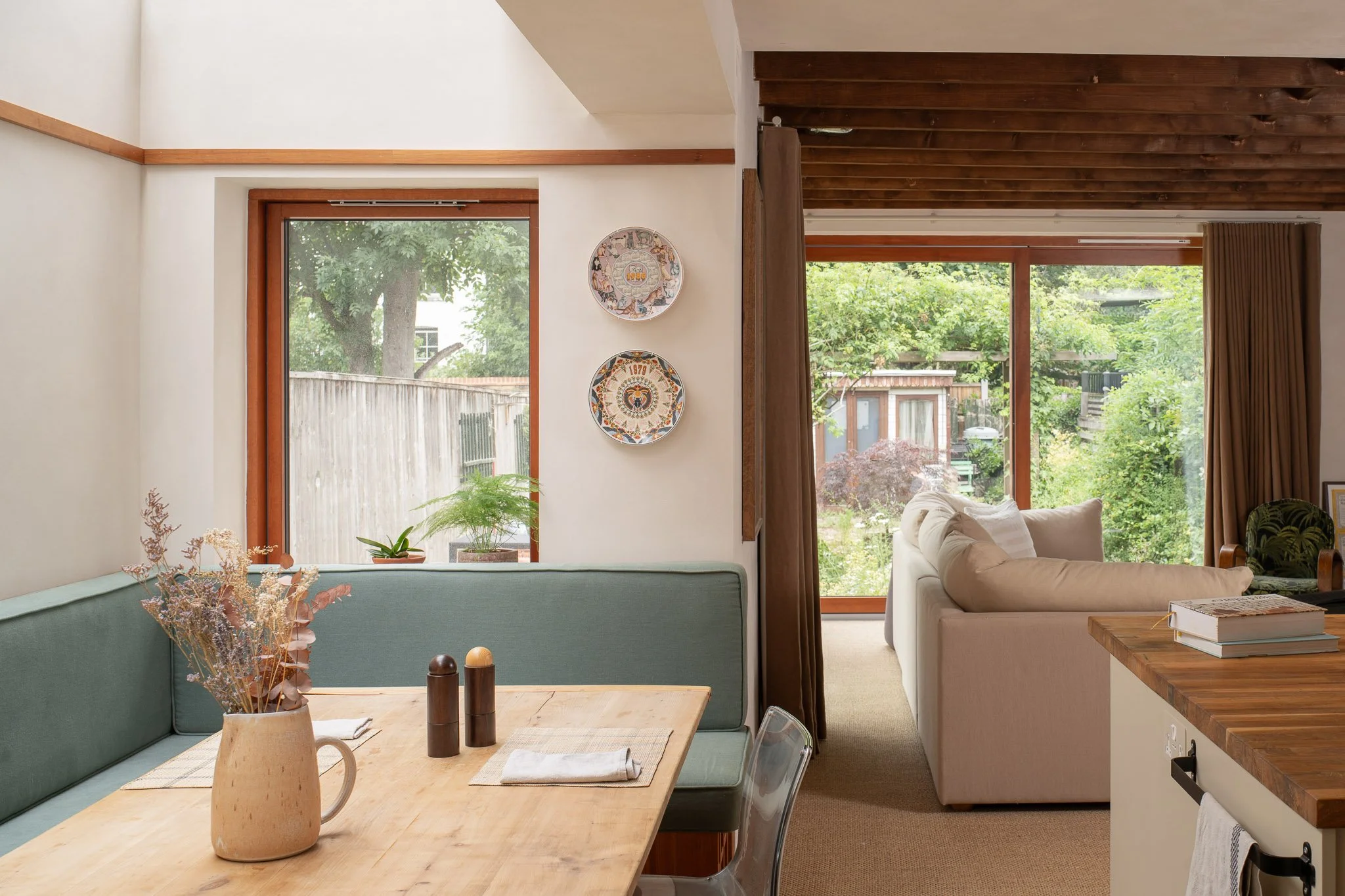 Interior of a living room with large windows showing greenery outside, beige sofas, wooden ceiling, and decorative plates on the wall.