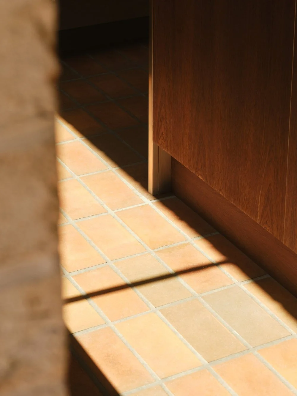 Sunlit kitchen floor with terracotta tiles, part of wooden cabinet, and shadows cast on the tiles.