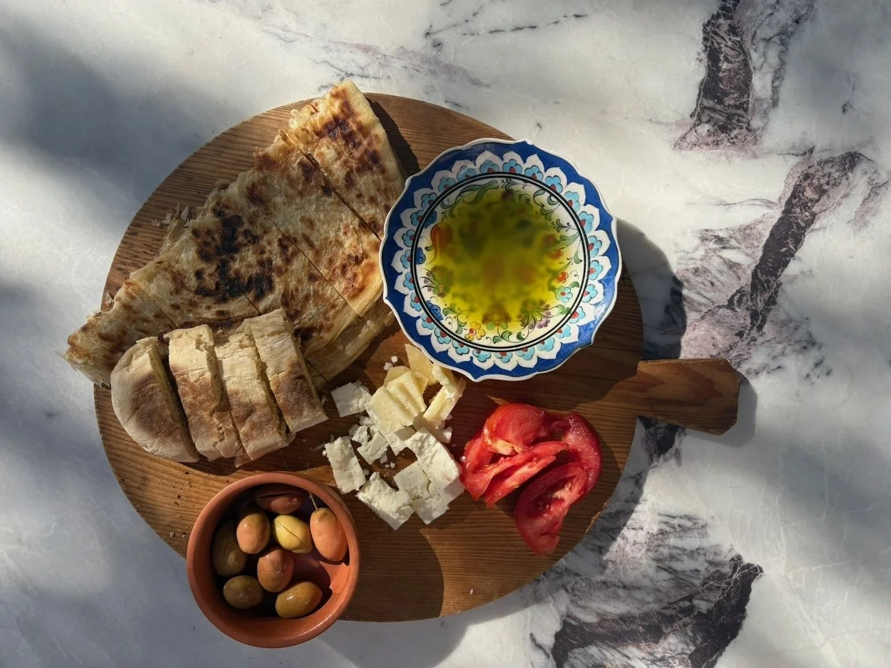 A wooden cutting board with slices of flatbread, a small bowl of green and yellow oil or sauce, chopped white cheese, slices of tomato, and a small bowl of green and black olives on a marble surface.
