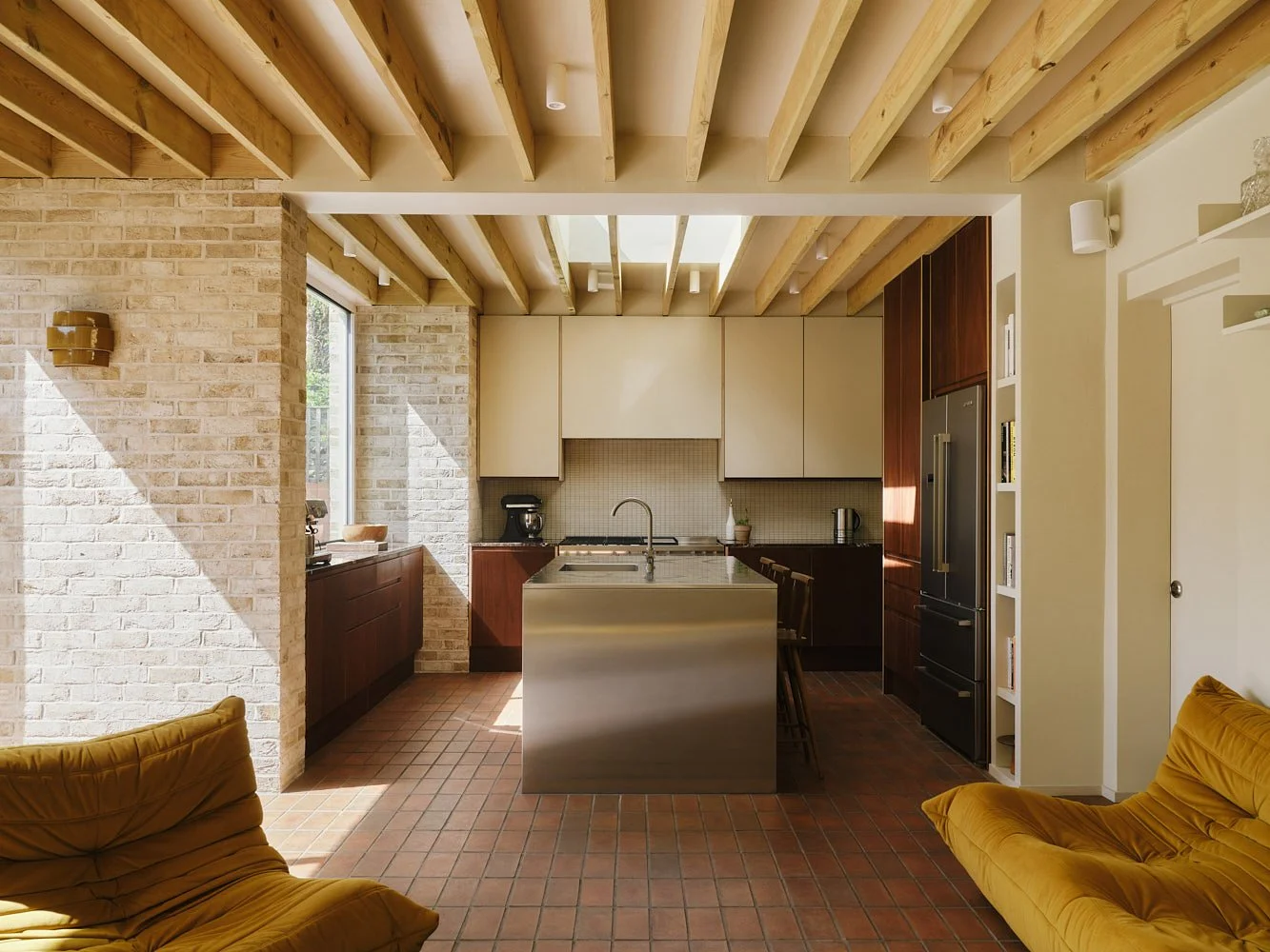 Open-concept kitchen and living room with brick walls, exposed wooden beams, and yellow armchairs