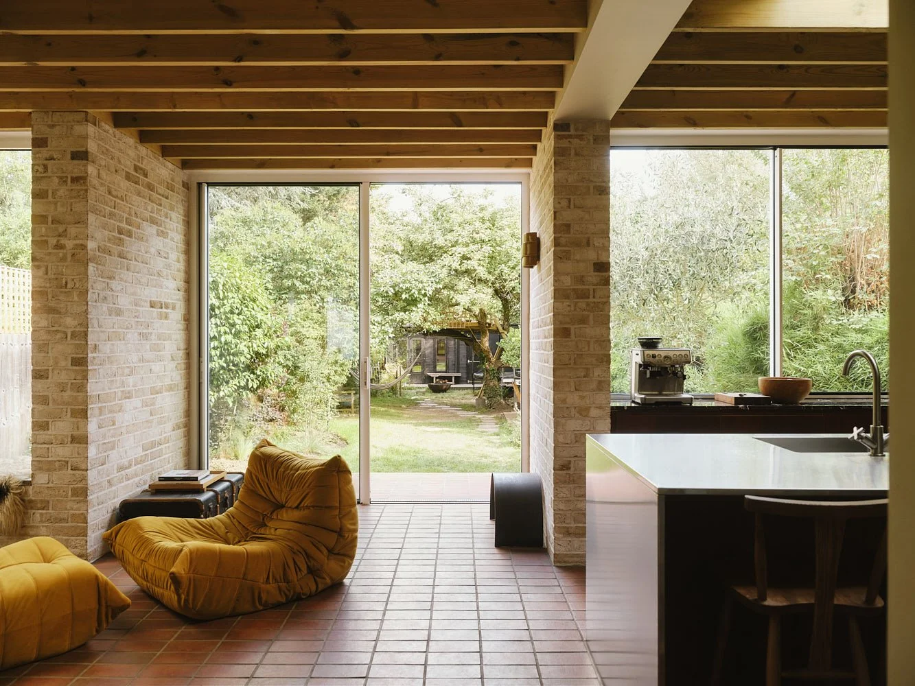 Bright living room with large windows showing an outdoor garden, yellow lounge chair, matching ottoman, kitchen with coffee machine, bowl, and sink.