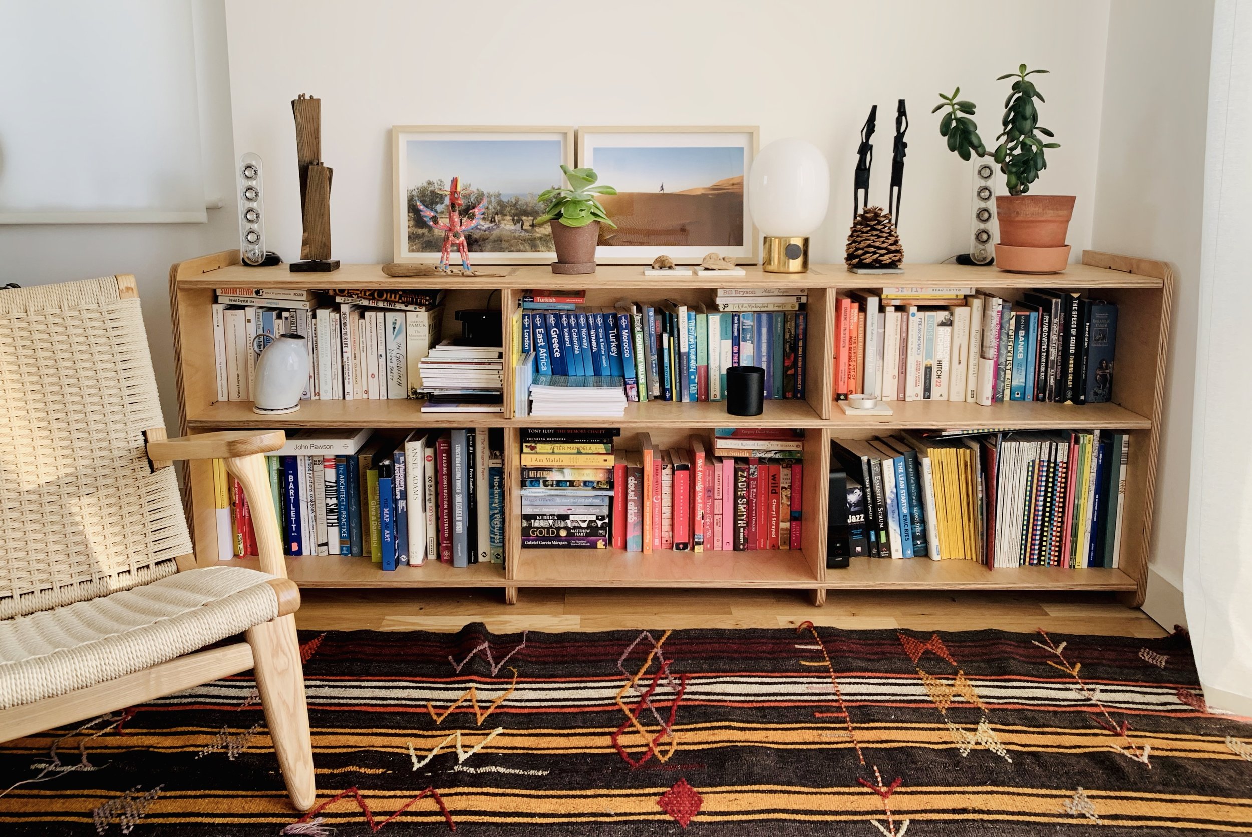 A wooden bookshelf filled with books and decorative items, topped with framed landscape photographs, potted plants, and sculptures. A wicker chair and a colorful rug are partially visible in the foreground.