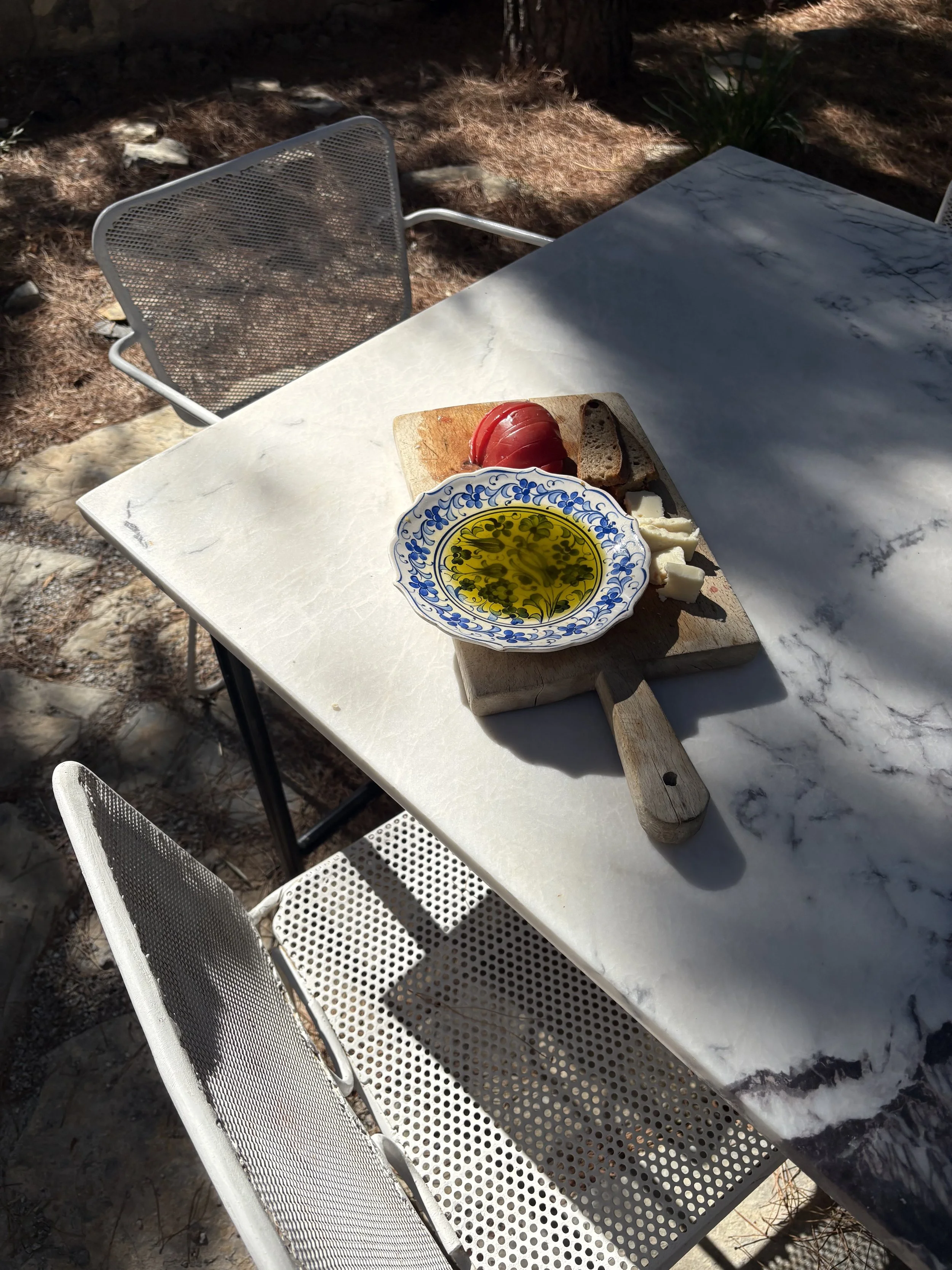 A white outdoor table with a marble pattern, set in a shadowy garden area, with a chair on one side and a plate of food including sliced cheese, tomato, bread, and olive oil on a cutting board.