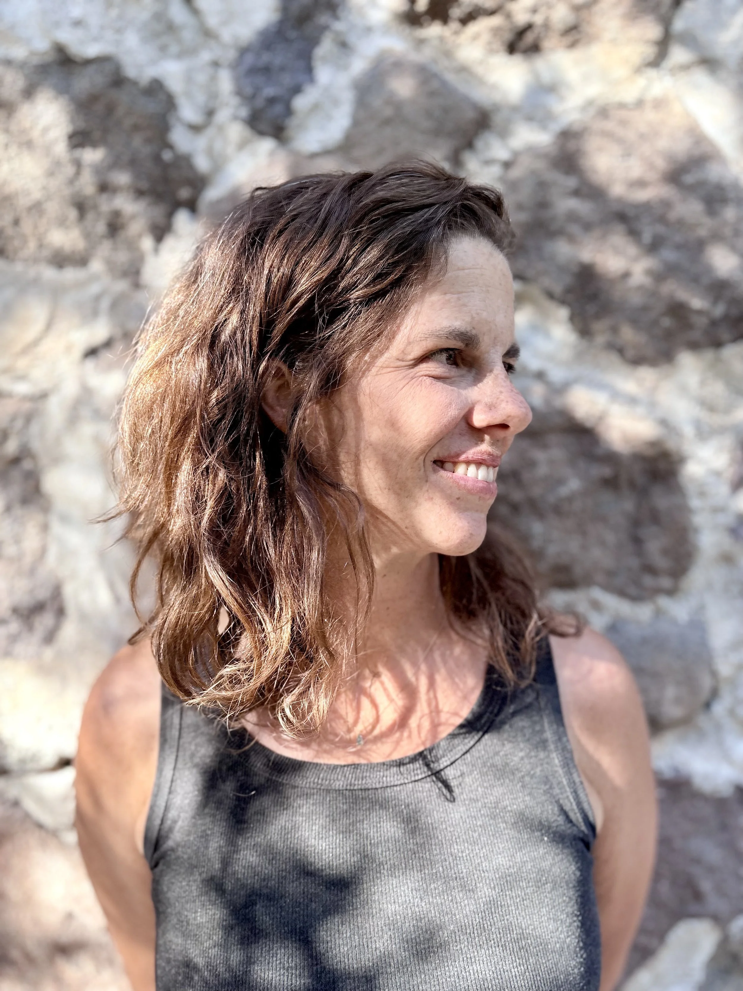 Woman with long, wavy brown hair smiling outdoors near a rocky wall.