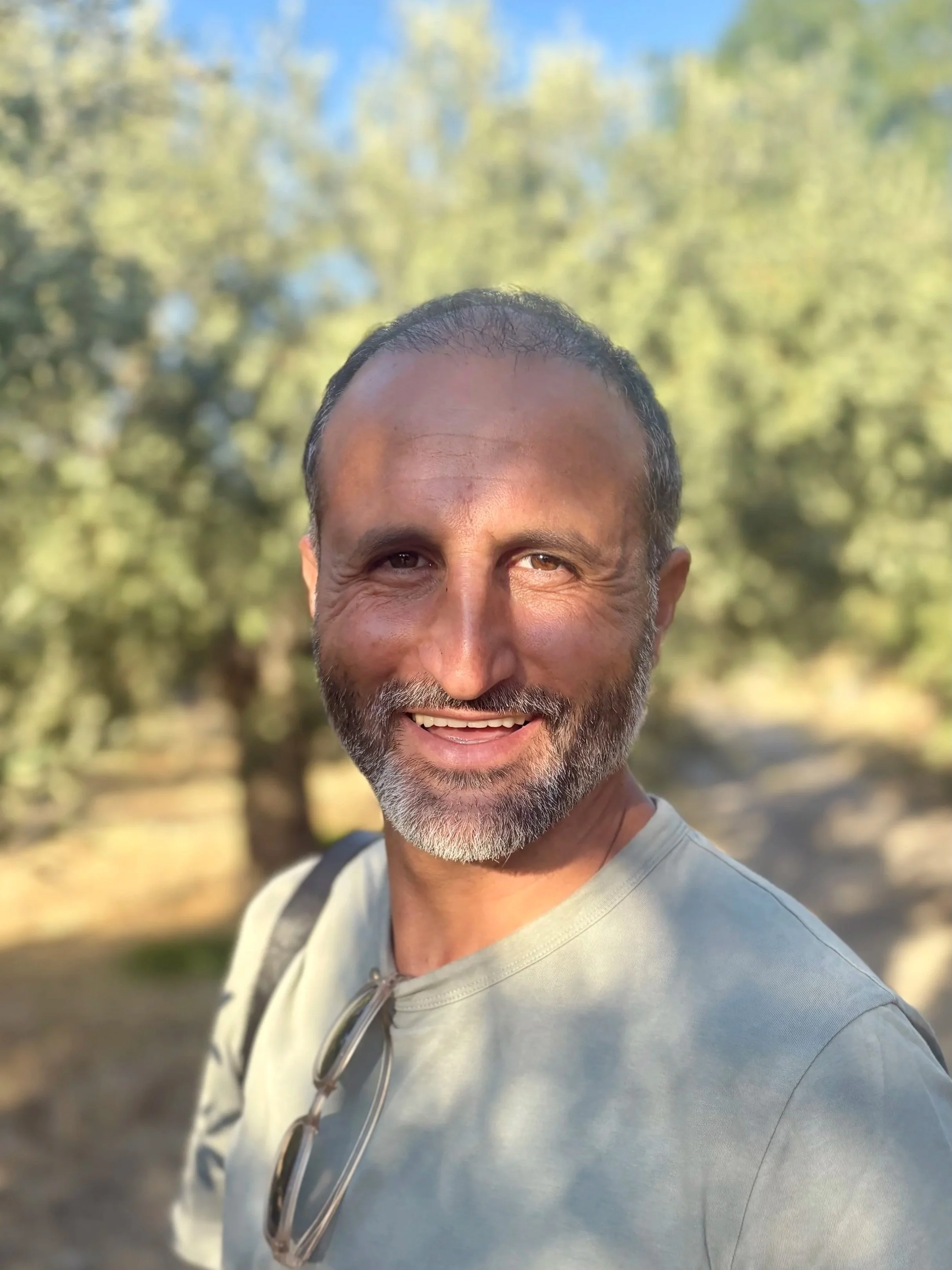 A smiling man with short dark hair and a beard, wearing a light-colored shirt, outdoors with blurred trees and sky in the background.