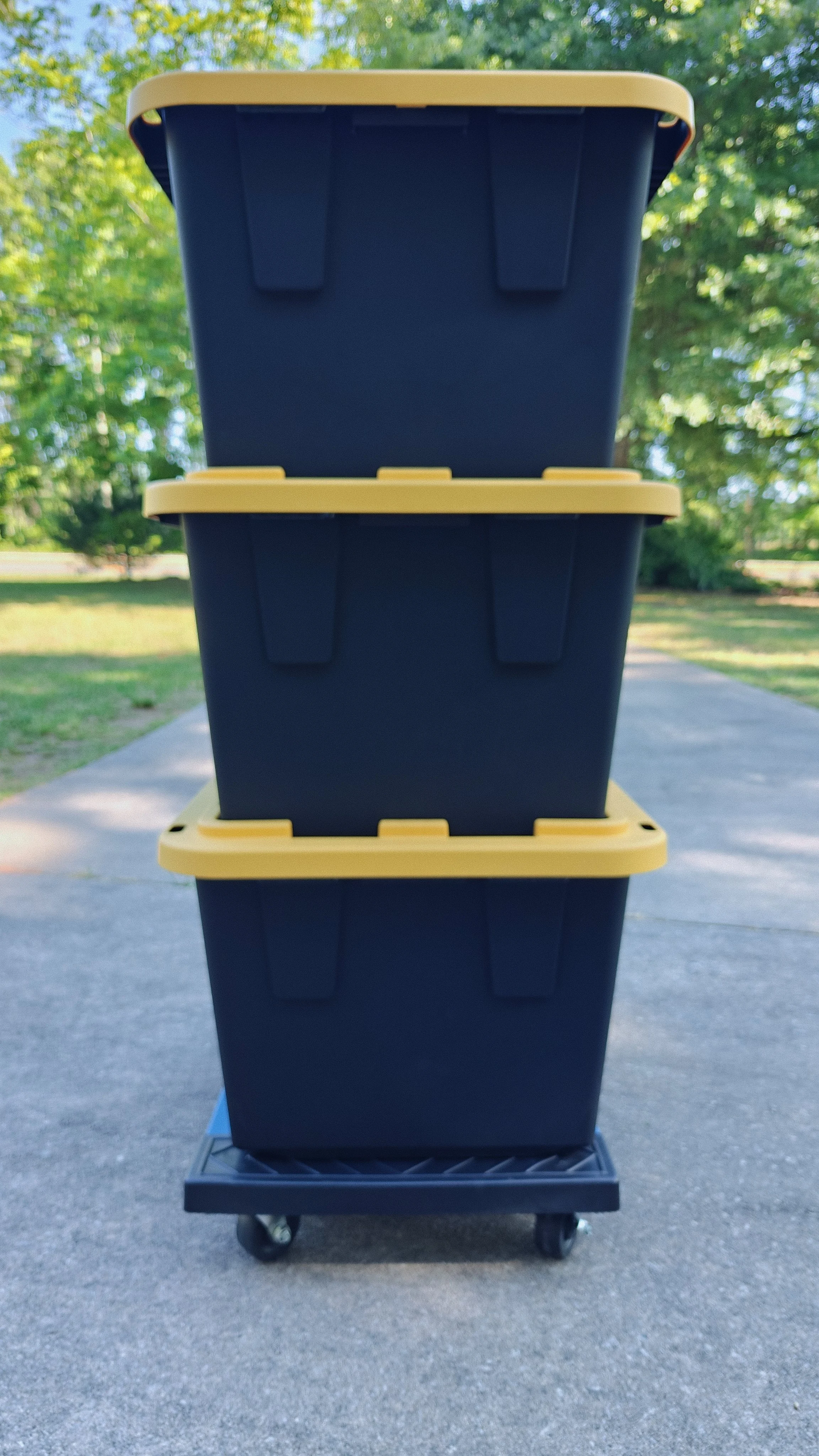 Stacked blue and yellow plastic storage containers on a rolling cart outdoors with trees in the background.