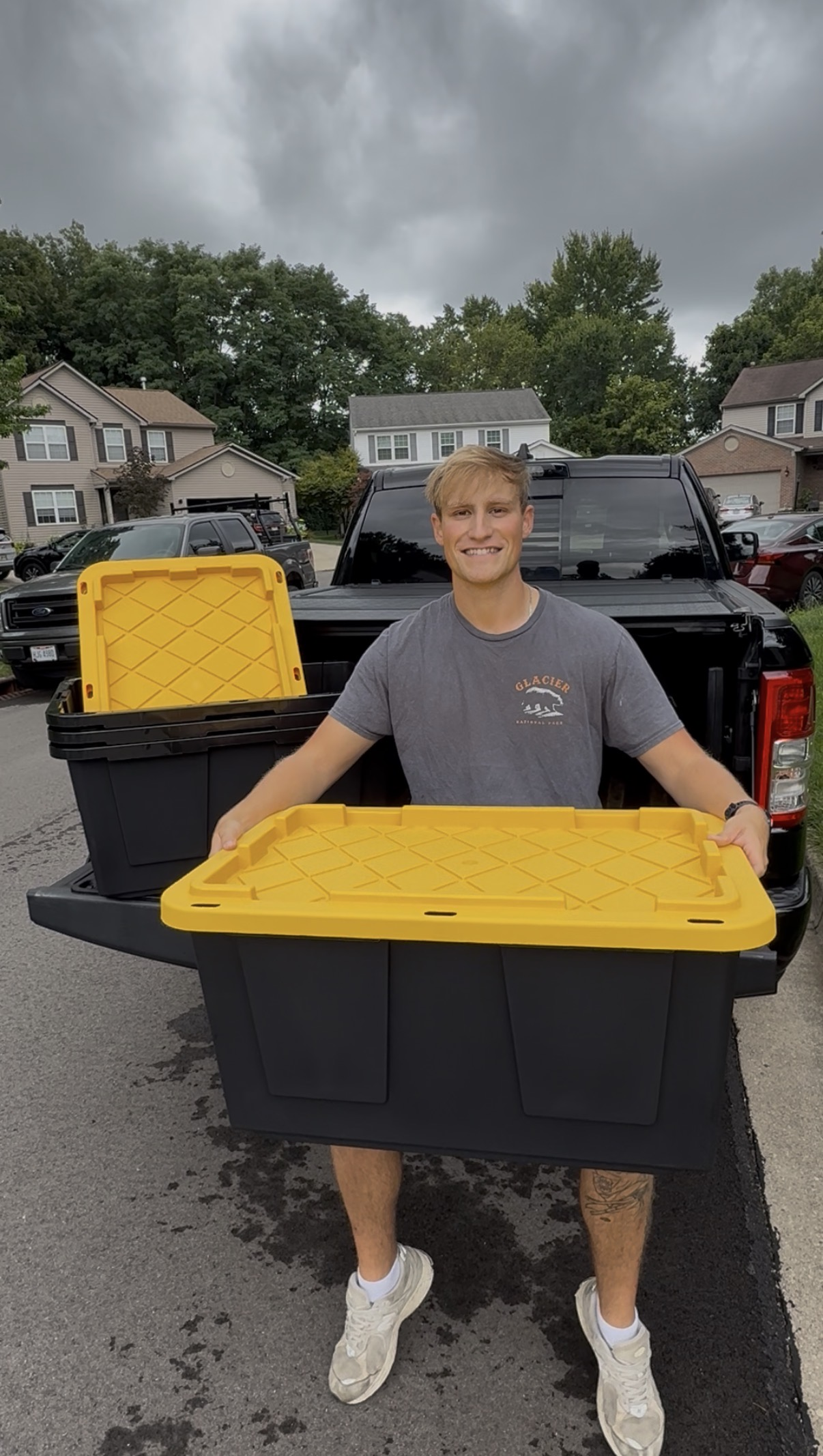 A young man smiling and holding a large plastic moving box in front of a black pickup truck parked in a suburban neighborhood. Moving boxes for rent.