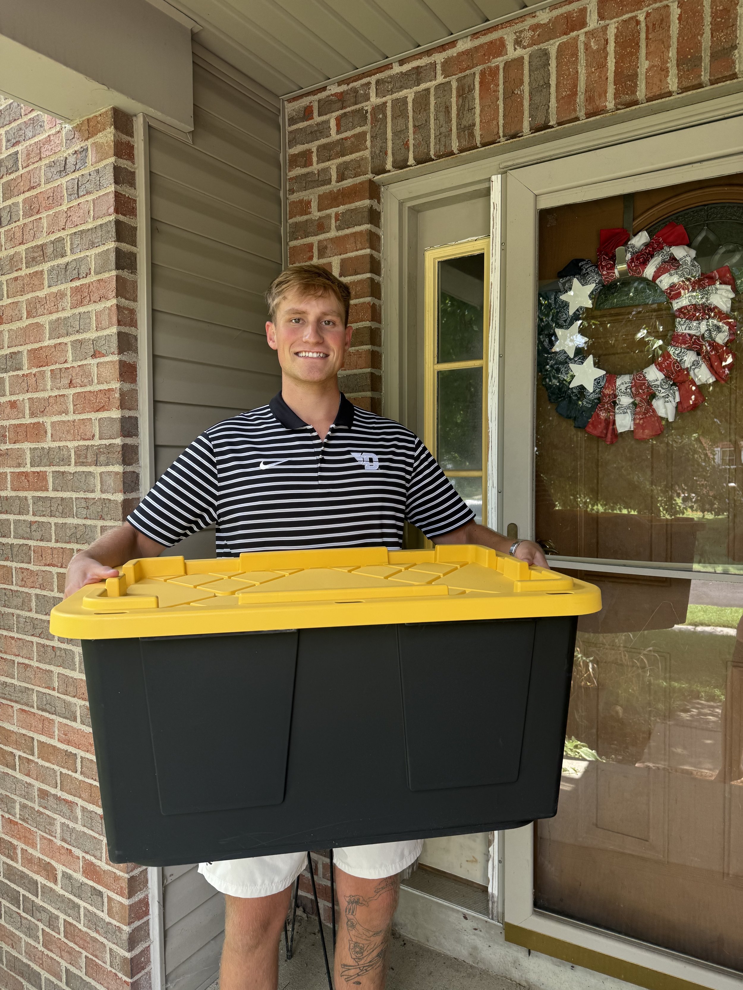 Person standing at a door holding a black and yellow storage bin, wearing a black and white striped shirt and white shorts, with tattooed legs, in front of a brick and siding house with a wreath on the door.