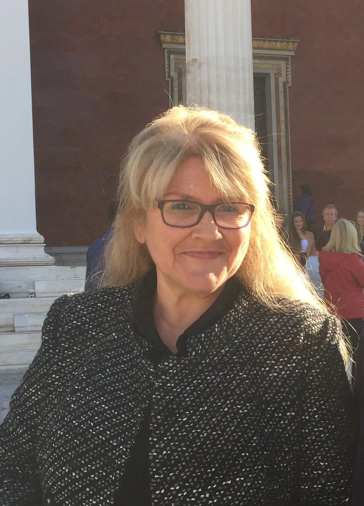 A woman with blonde hair, glasses, and a black and white patterned coat smiling at the camera outdoors with a historic building and steps in the background.