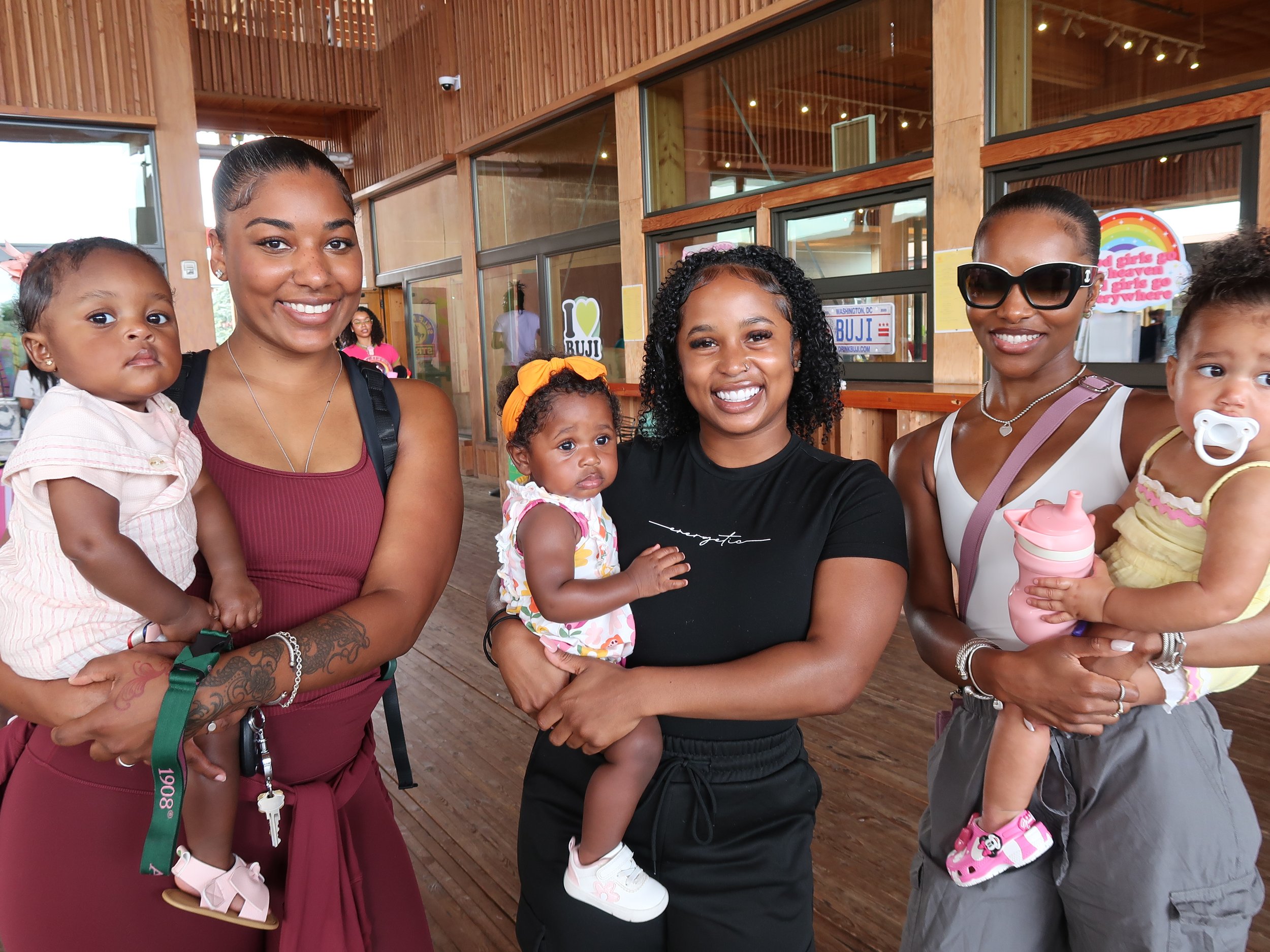 Group of women holding children indoors with wooden interior and social media signs in the background.