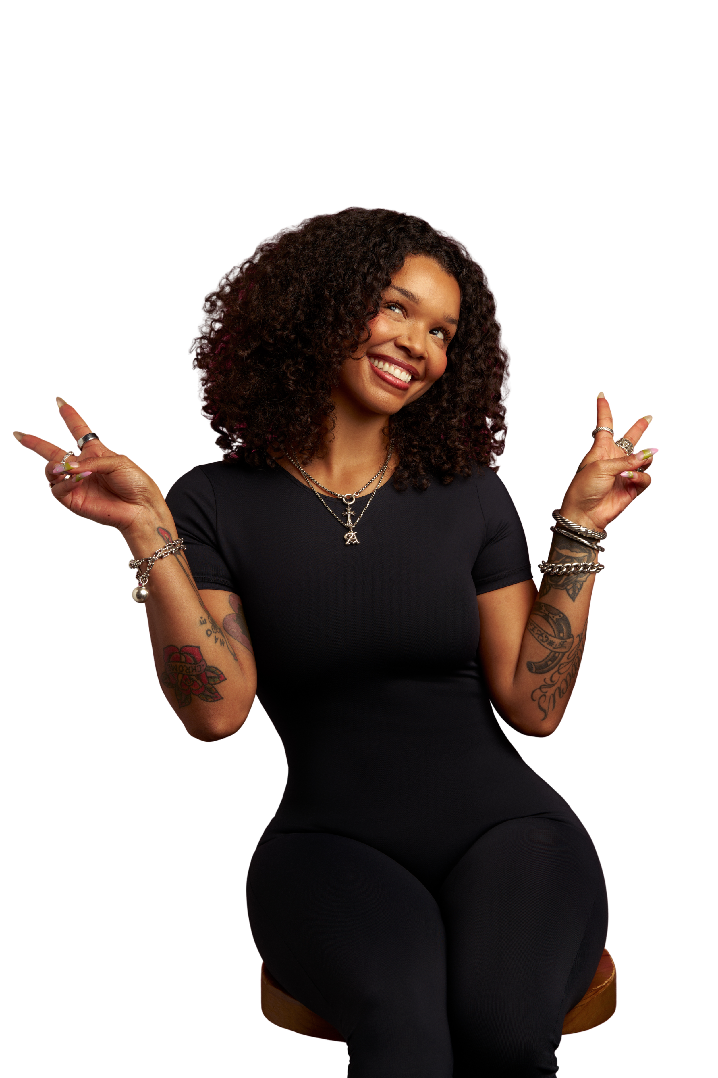 A woman with curly hair smiling, sitting on a stool, making peace signs with both hands, wearing a black t-shirt and jewelry, with tattoos on her arms, against a black background.