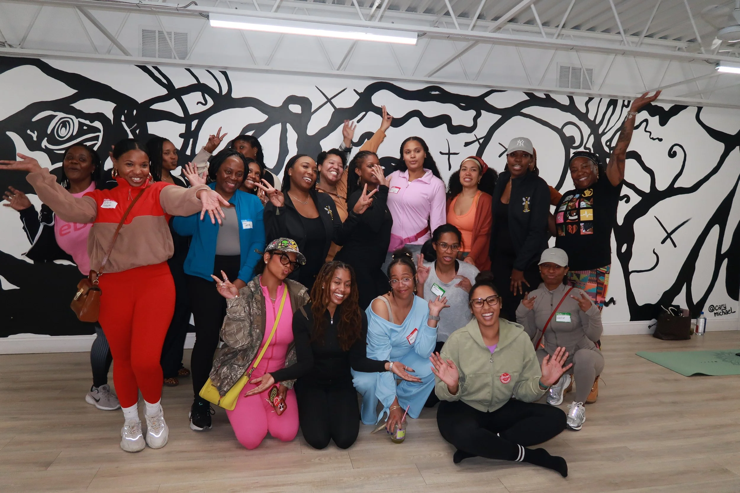 Group of diverse women and a few young girls posing inside a building with a large black and white mural of a tree on the wall behind them. They are smiling, making peace signs, and wearing casual and colorful clothing.