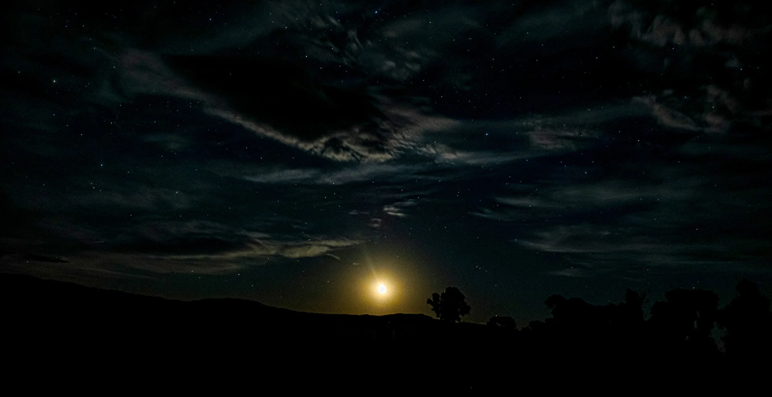 Moon illuminating dramatic clouds above the Wyoming landscape beneath a star-filled night sky.