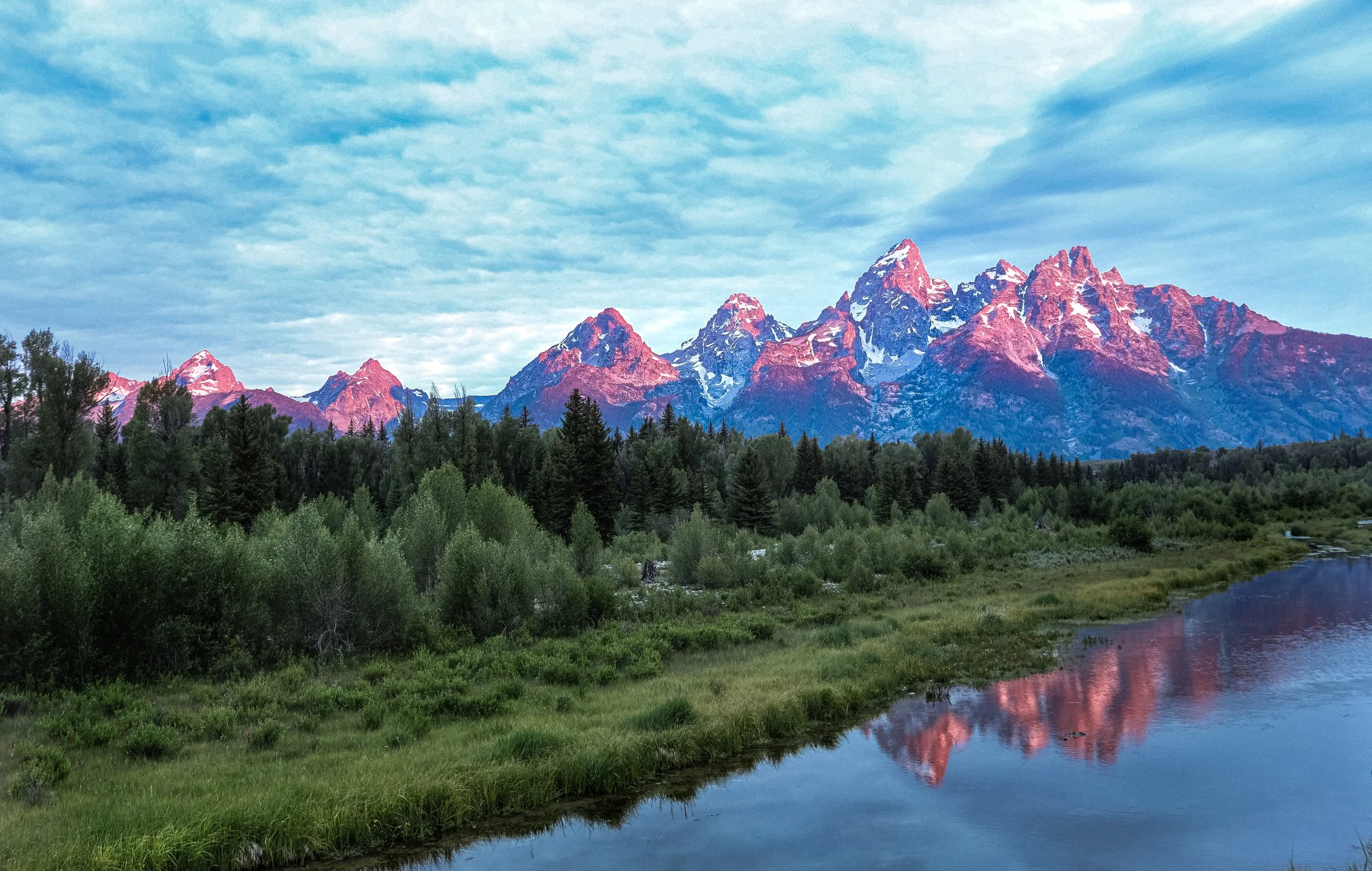 Mountain range with snow on peaks, forest in foreground, river reflecting mountains and sky