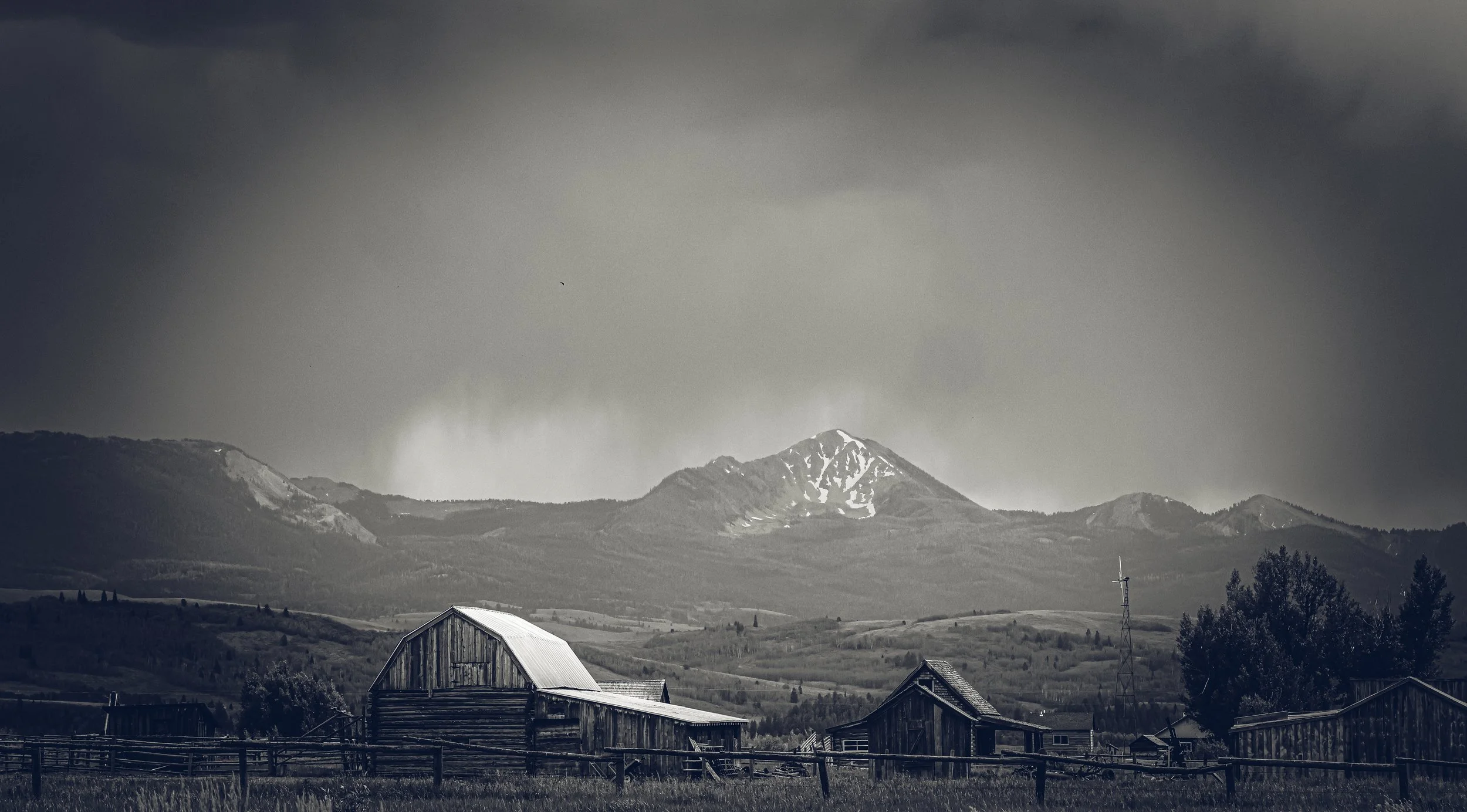 Black and white landscape photograph of Mormon Row barns beneath the Teton Range in Grand Teton National Park, Wyoming, with dramatic storm clouds surrounding snow-capped peaks.
