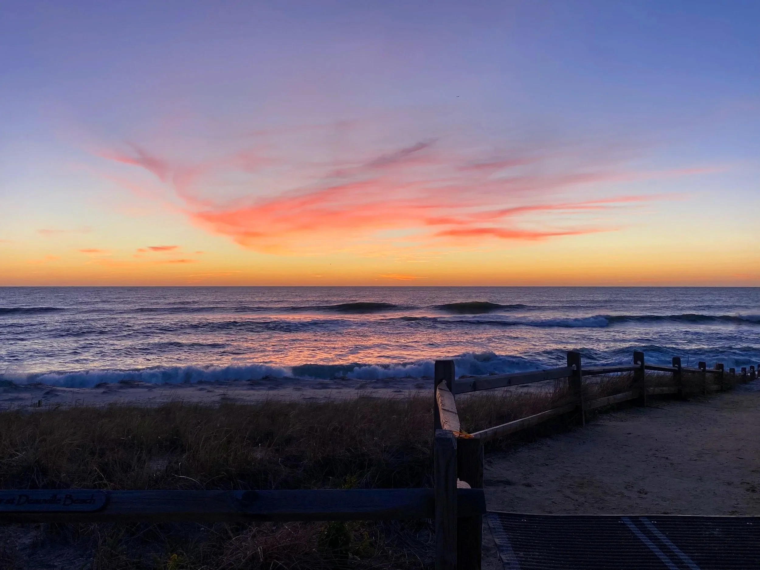 Sunset over the ocean with colorful pink, orange, and purple sky and gentle waves. A wooden fence in the foreground along a sandy beach.