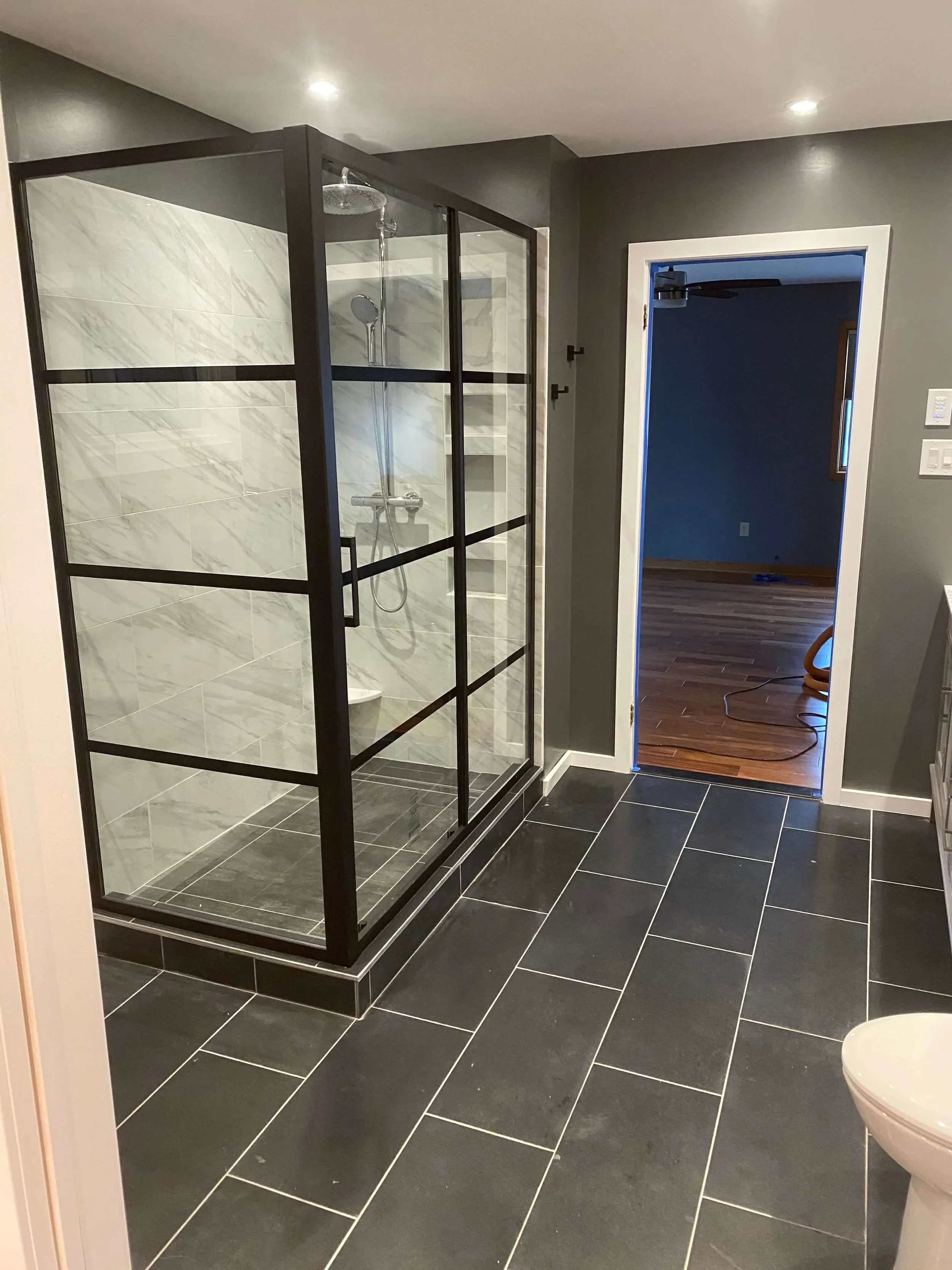 Modern bathroom with black tiled floor, glass shower enclosure with marble tile walls, and a doorway leading to another room with wood flooring and blue walls.