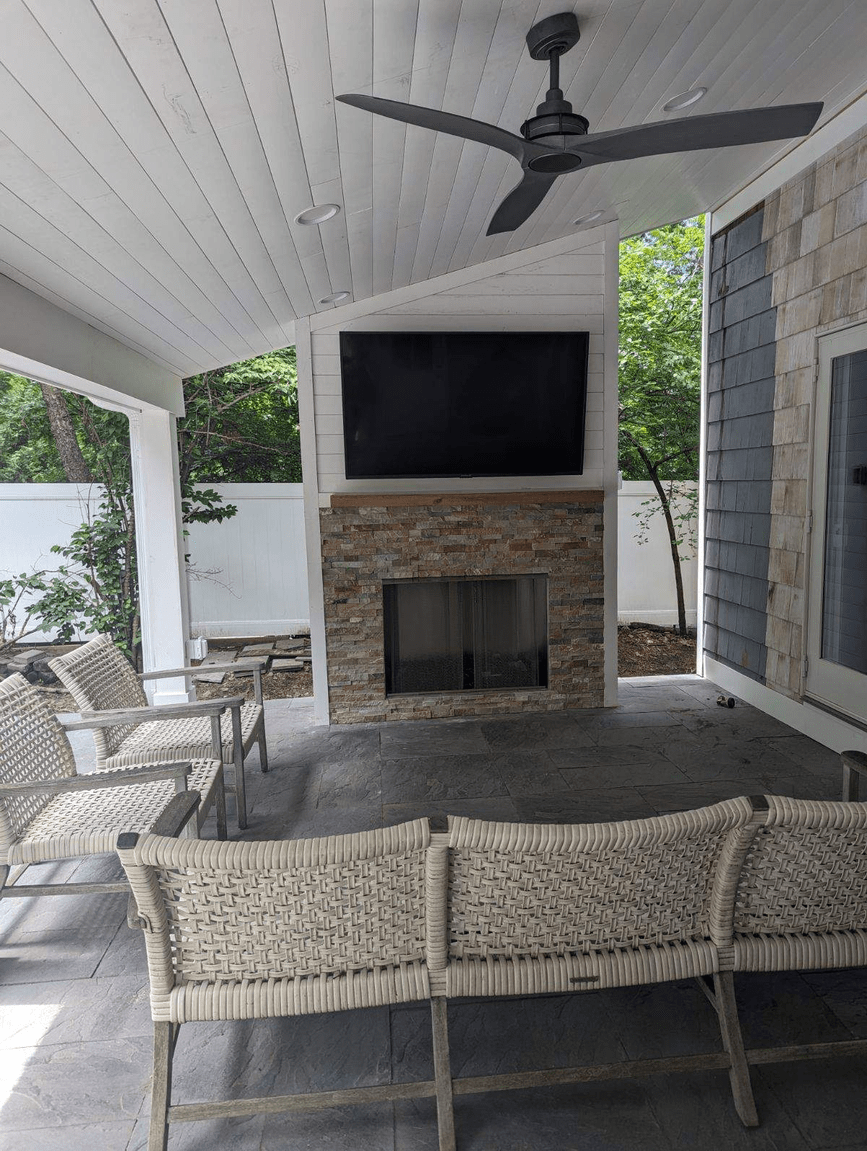 Covered outdoor patio with a ceiling fan, a flat-screen TV above a stone fireplace, wicker chairs, and a stone floor, with trees in the background.