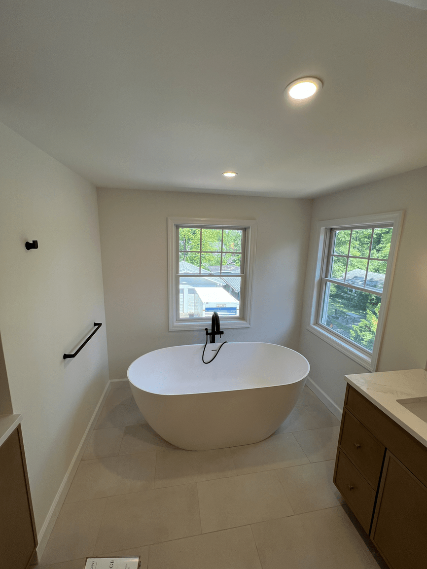 Modern bathroom with a freestanding bathtub, two large windows, a beige vanity, and recessed ceiling lights.
