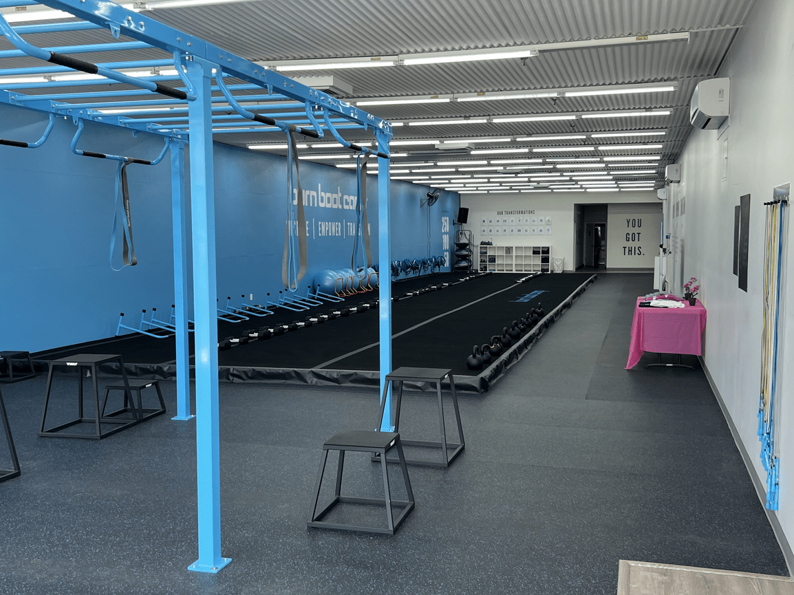 Interior of a fitness gym with black rubber flooring, blue workout equipment, kettlebells on a raised platform, shelves, and motivational signs on the wall.