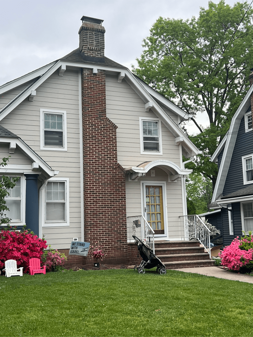 Front yard of a house with a brick chimney, surrounded by flowering plants, a stroller, and lawn chairs, with a "We Stand With Israel" sign.