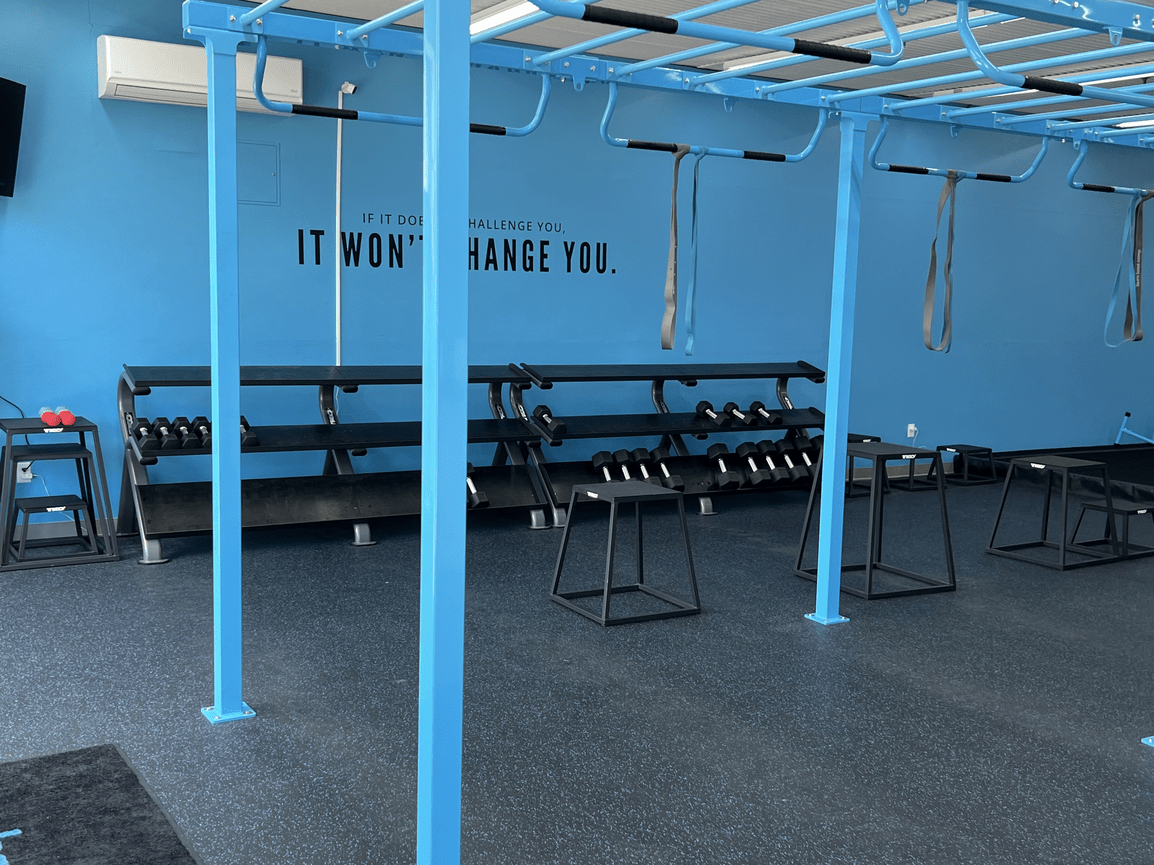 Inside a fitness gym with blue workout equipment, including pull-up bars, parallel bars, and weights. The wall has an inspirational quote: 'If it does not challenge you, it won't change you.'