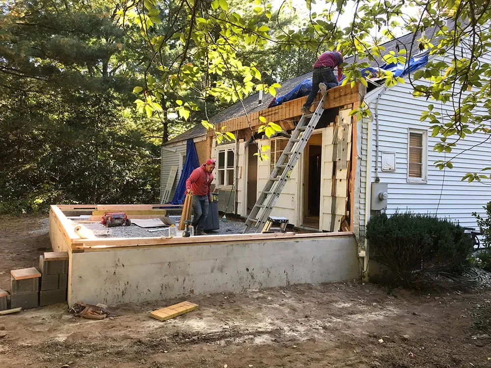 Construction workers building a new porch or extension on a house, with one worker on a ladder working on the roof and two others on the ground managing materials and tools, surrounded by trees.