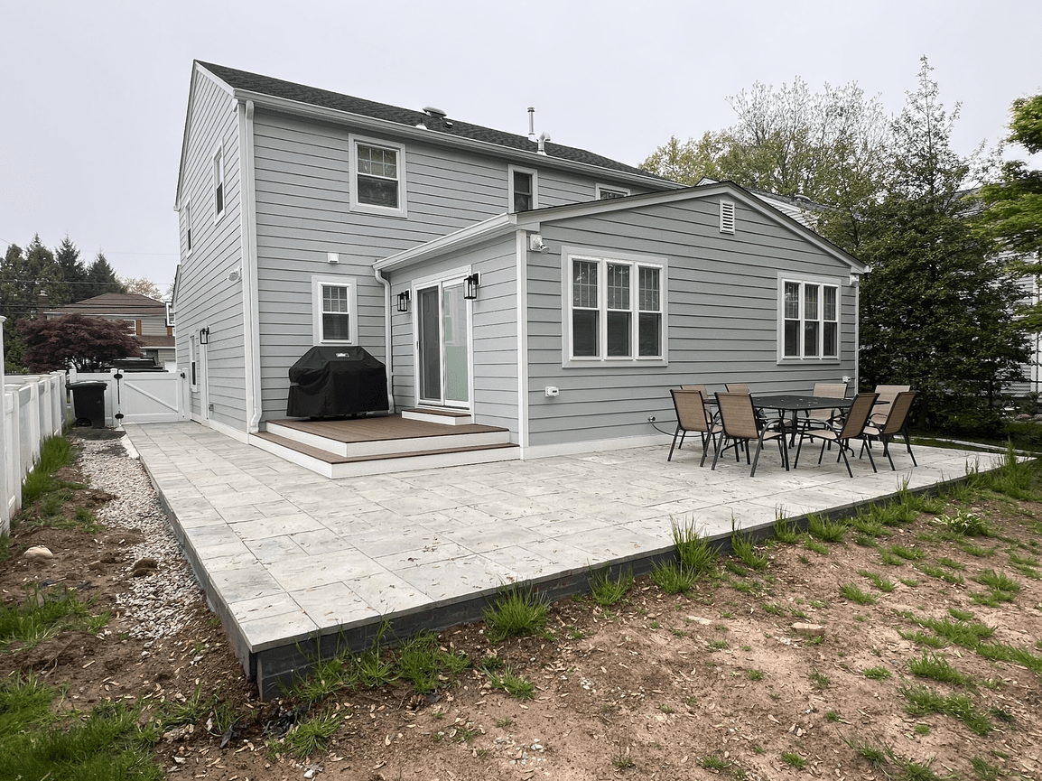 Backyard patio with outdoor dining table and chairs, patio with steps leading to a house with gray siding, white trim, and a sliding glass door, and a black grill, with trees in the background.
