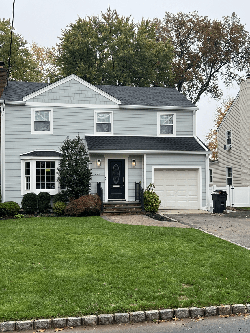 Light gray two-story house with black front door, white window frames, front porch with stairs, attached garage, and a well-maintained lawn with bushes and trees in the background.