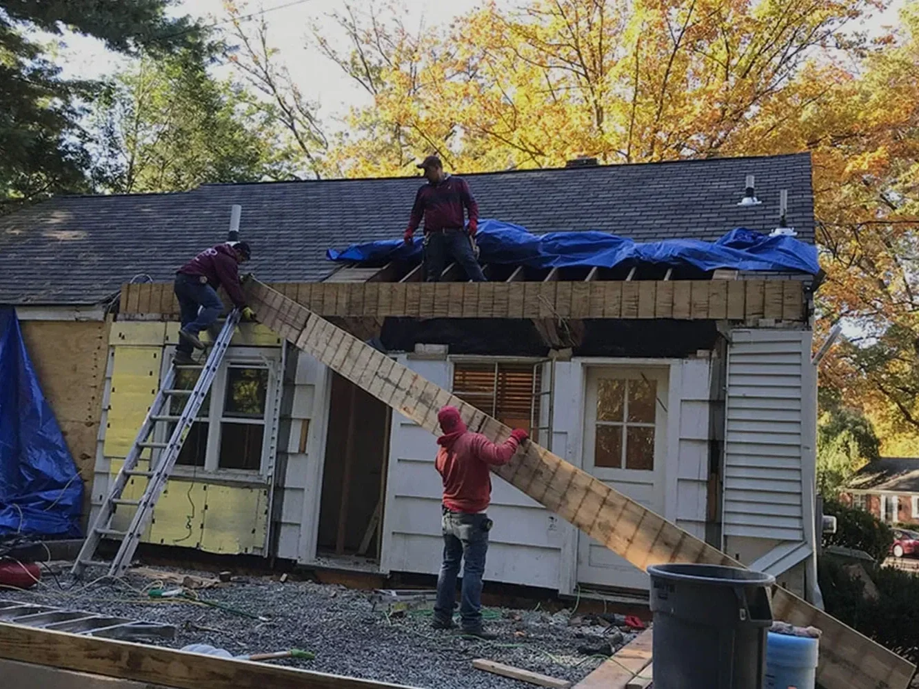 Construction workers rebuilding a house's roof, with wooden beams and blue tarps, in a neighborhood with trees in autumn.