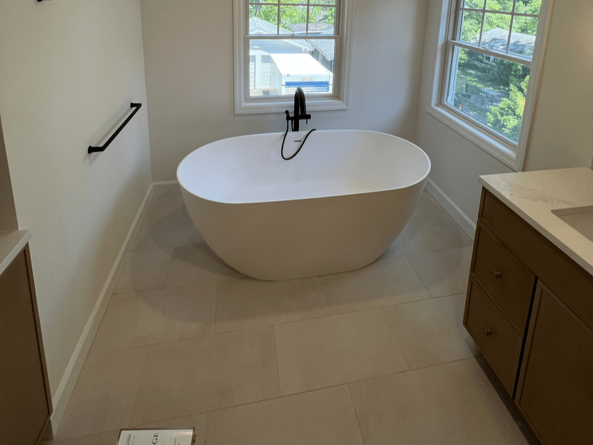 A modern bathroom featuring a white freestanding bathtub near two large windows, with a black faucet, a wooden cabinet, and a light-colored tiled floor.