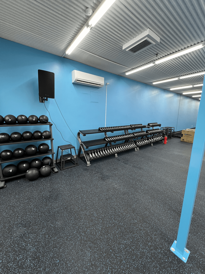 Interior of a gym room with blue walls, black rubber flooring, and ceiling lights. Equipment includes black medicine balls on a rack, a set of black dumbbells on a rack, a small black stool, and a red fire extinguisher. There is an air conditioning u