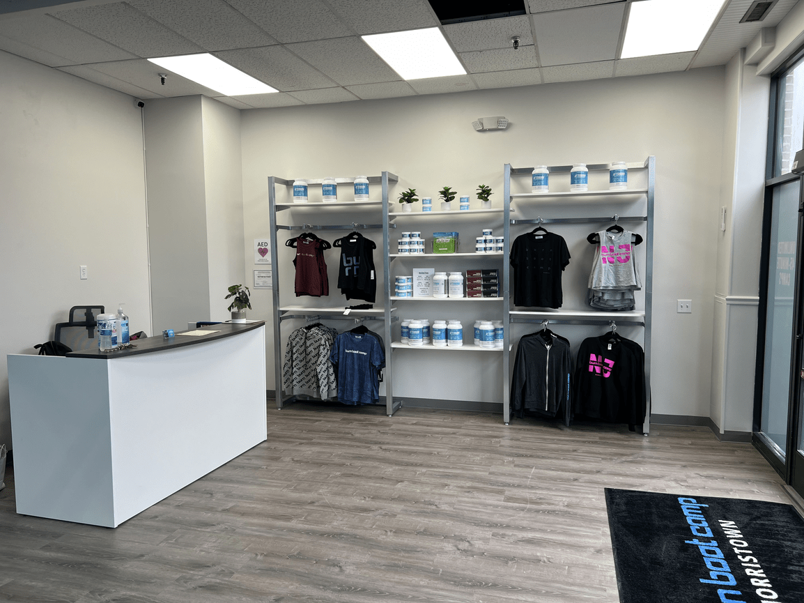 Inside a fitness supplement store with shelves of protein jars and athletic apparel, near a reception desk with water bottles, in a well-lit room with large window and wooden floor.