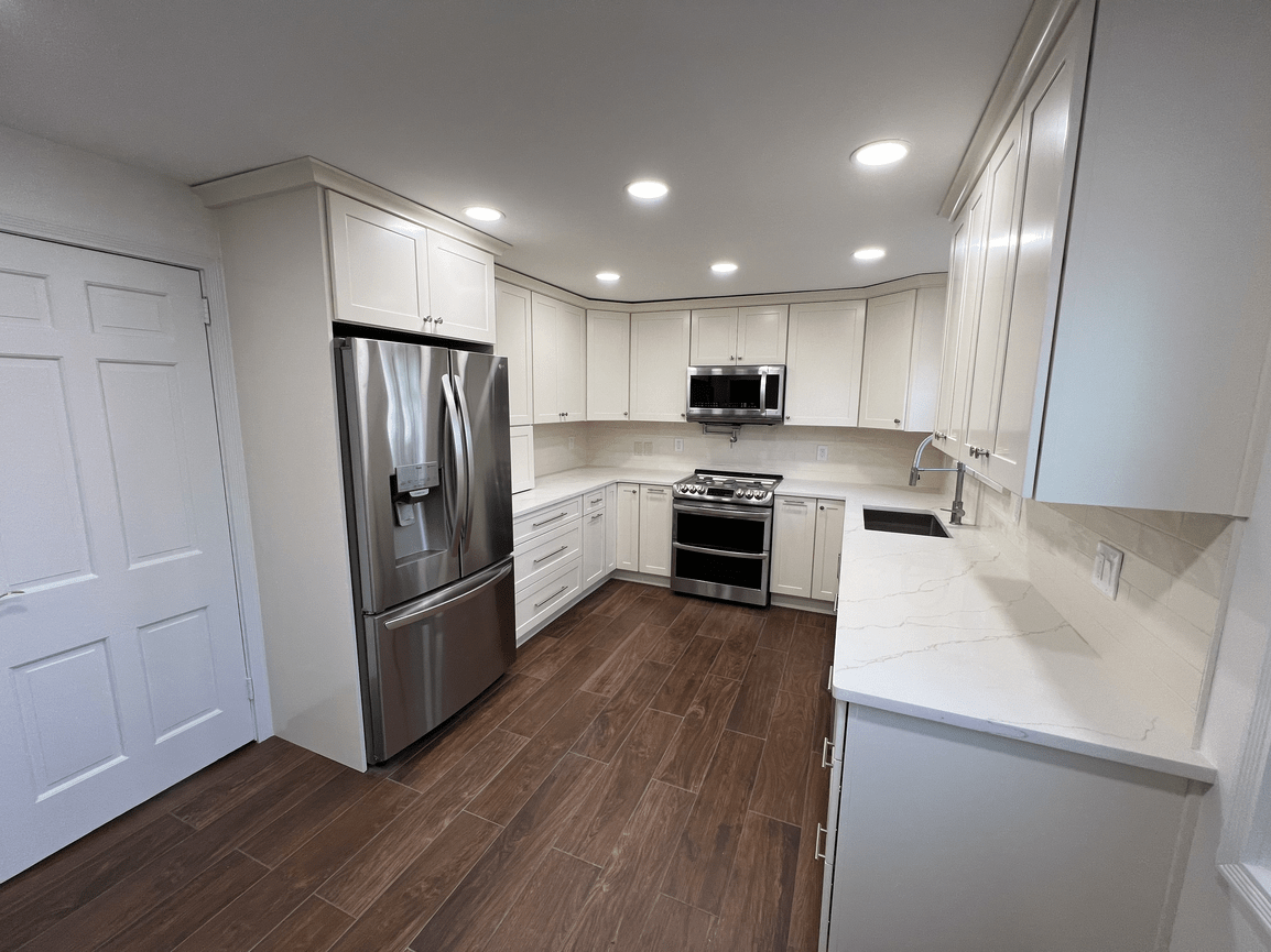 Kitchen with white cabinets, stainless steel refrigerator, microwave, and stove, brown wood-look flooring, and white countertops.