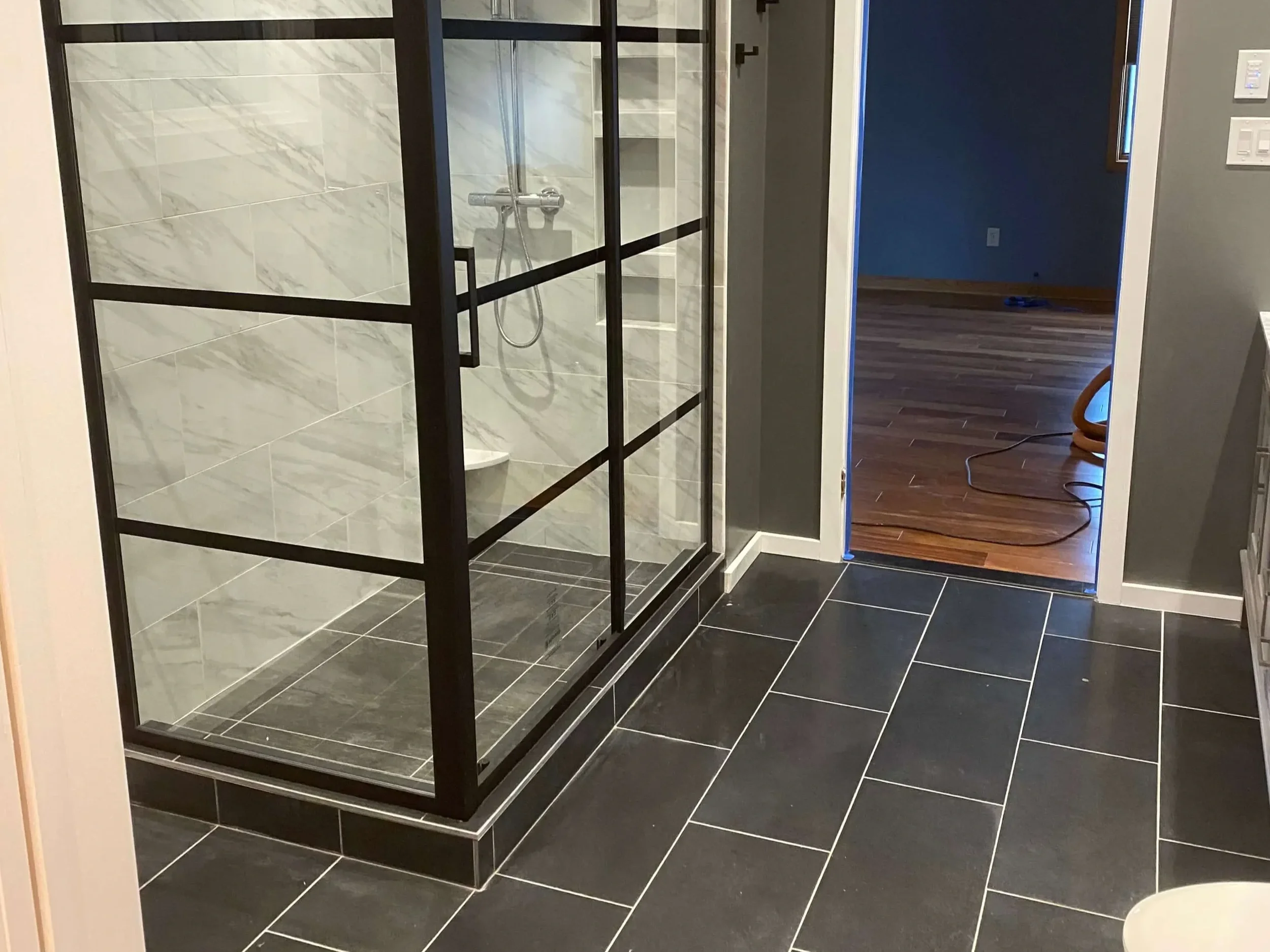 A modern bathroom with a walk-in shower enclosed by black-framed glass panels. The shower has marble-patterned tiles and a silver showerhead. The bathroom floor is tiled with large black tiles. To the right, a doorway leads to a room with hardwood flooring and a dark blue wall, with a coiled orange extension cord on the floor.