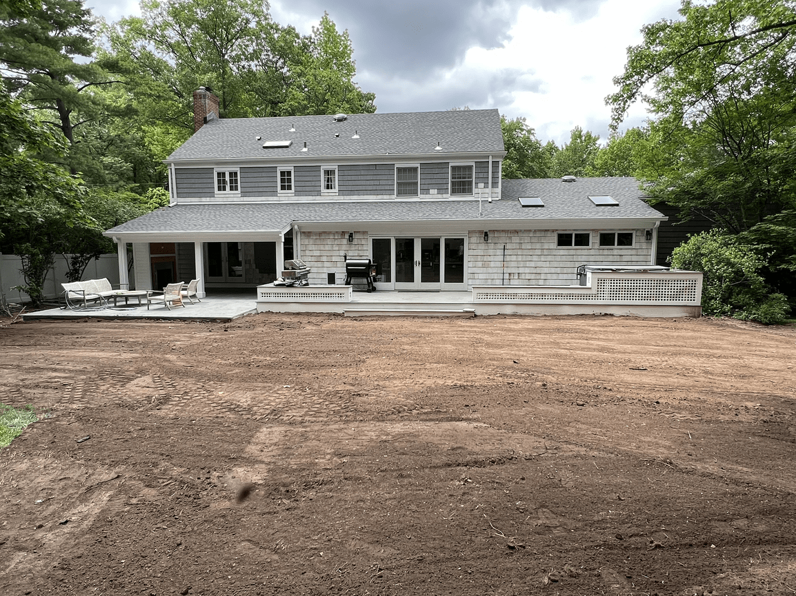 Backyard of a house with a patio, outdoor furniture, a barbecue grill, and trees surrounding the area, with a dirt ground in the foreground.