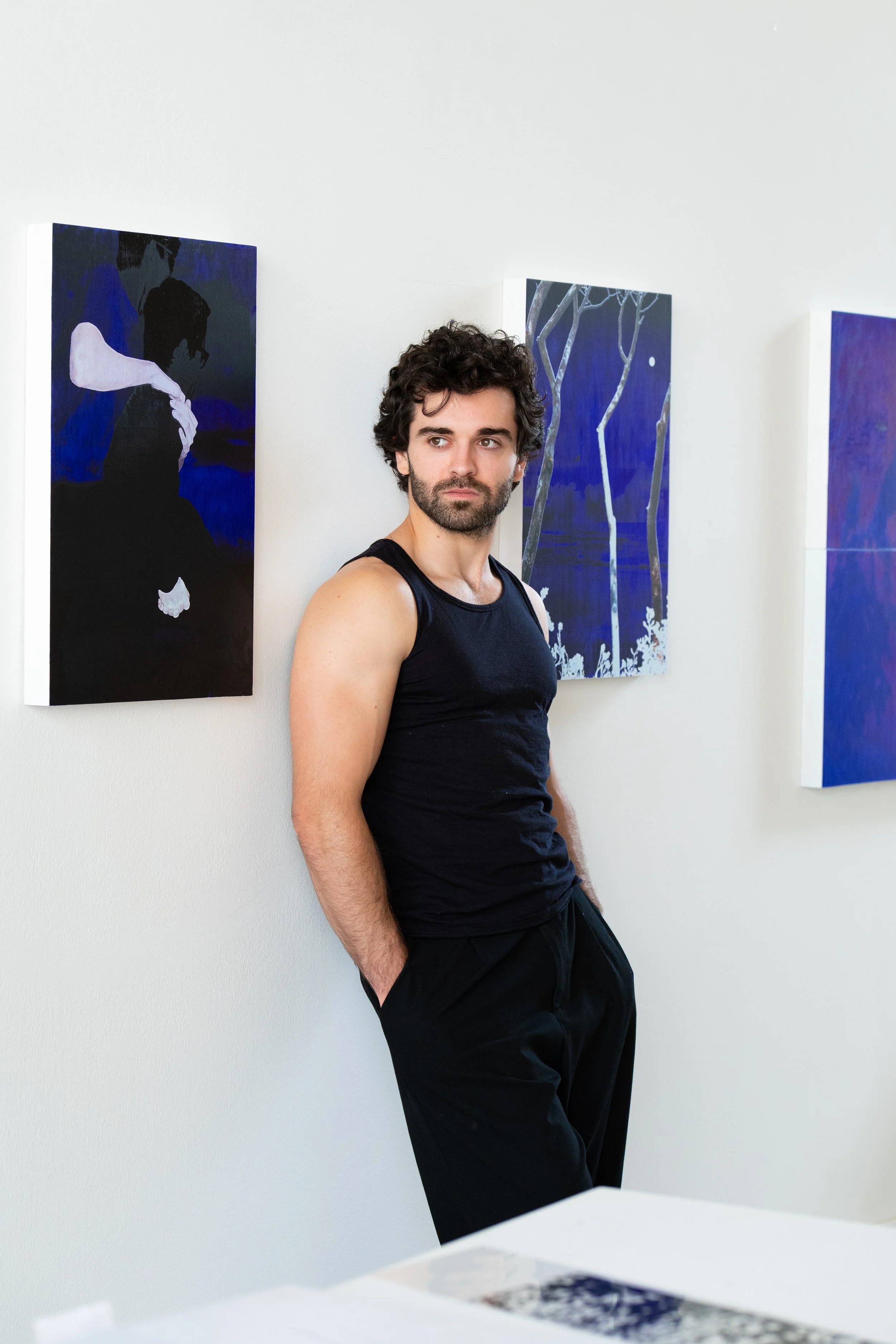 Dany Albiach, homme avec des cheveux ondulés et une barbe se tient dans une galerie d'art, portant un t-shirt noir et un pantalon noir, debout près de trois tableaux abstraits avec des tons de bleu et de noir.