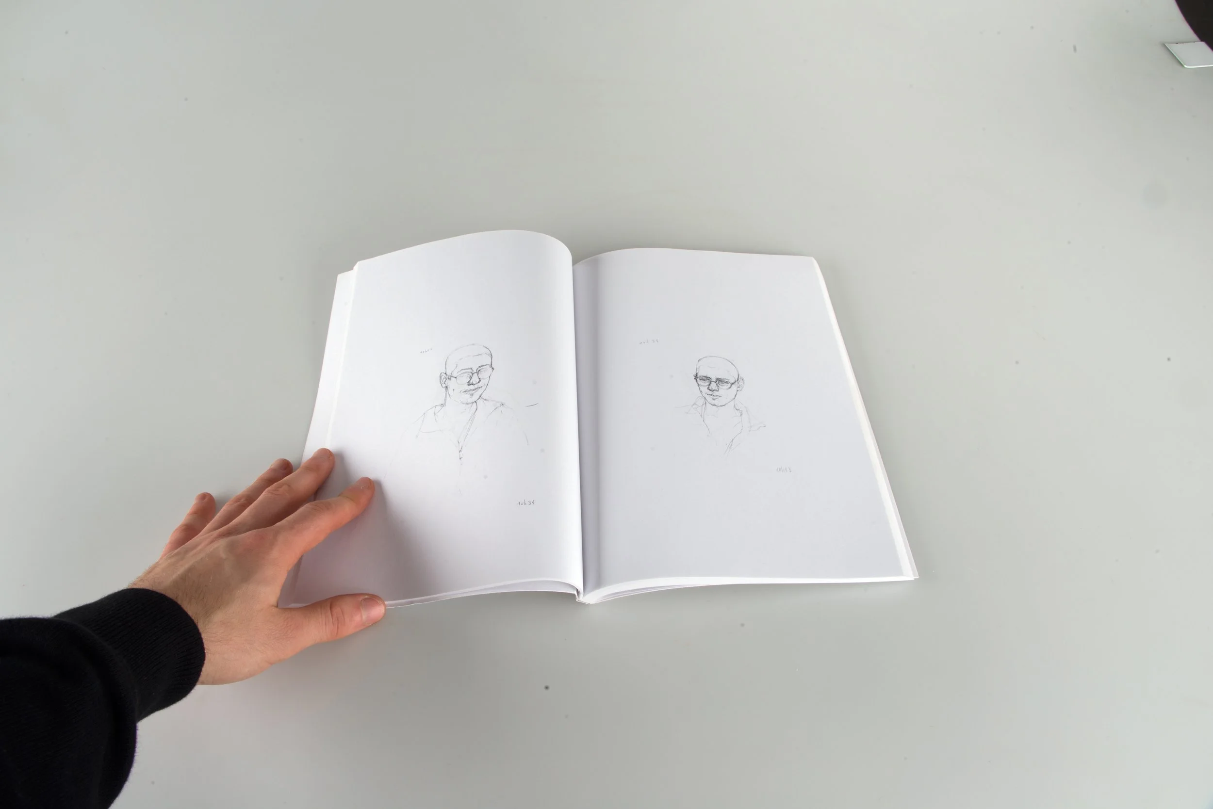 Une main tient un carnet ouvert avec des dessins de portraits de personnes portant des lunettes, vues de face.