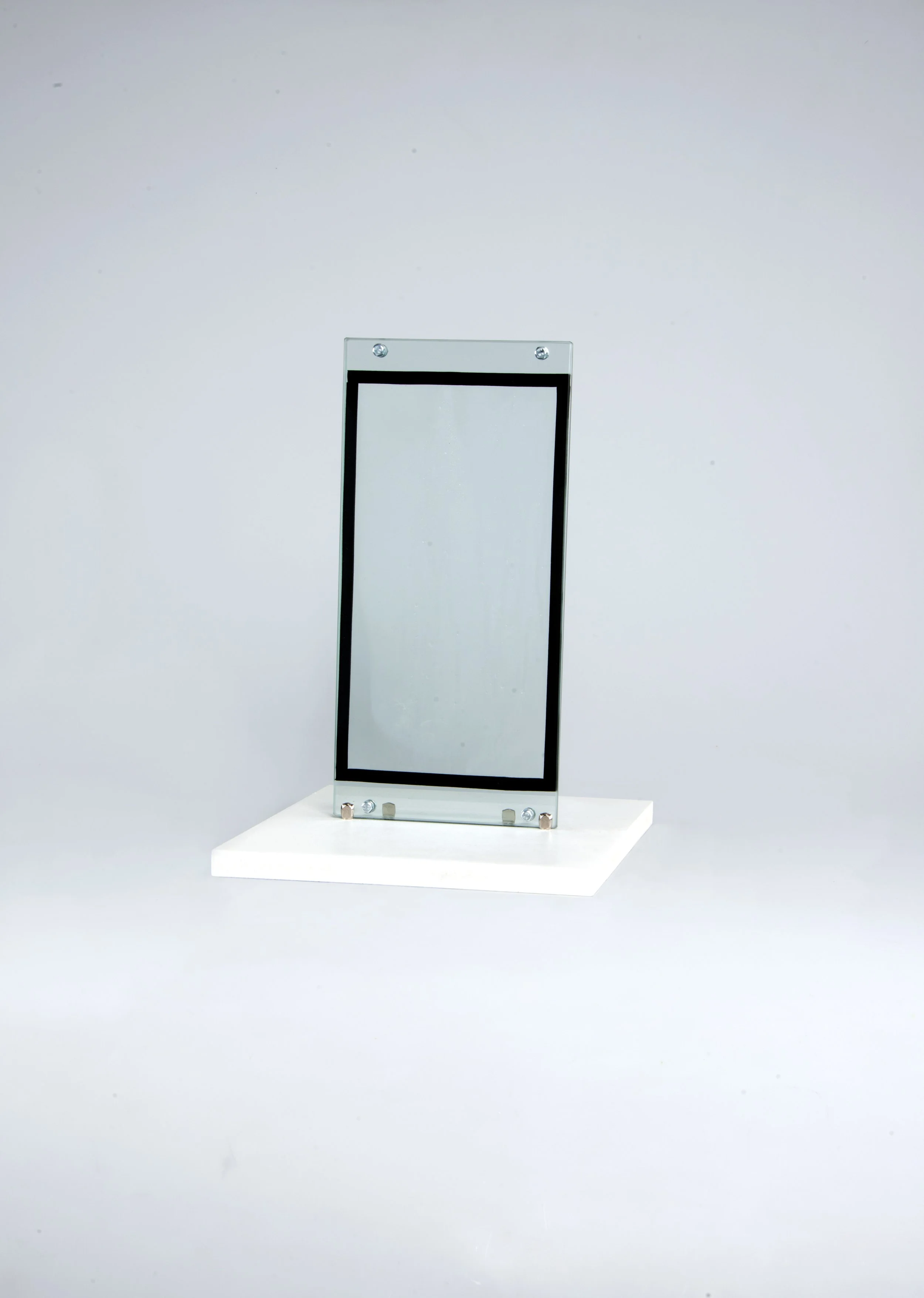 Une structure en verre transparent avec un cadre noir au centre, fixée à une base blanche, sur un fond gris clair.