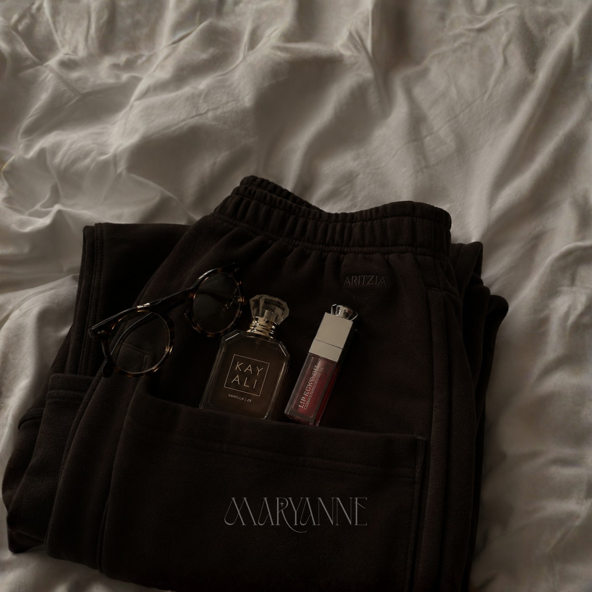 A crumpled white bedsheet with black sweatpants lying on top, with sunglasses and a perfume bottle placed on the sweatpants.
