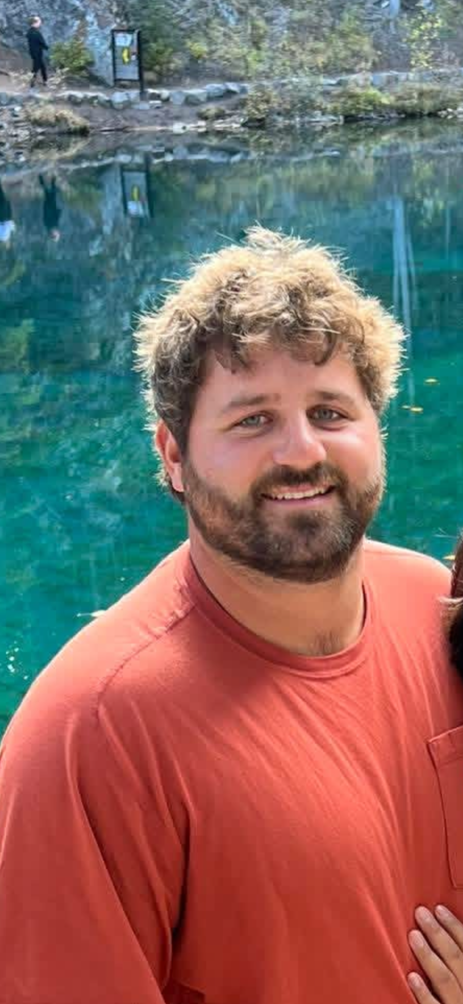 A smiling man with curly hair and a beard wearing a red shirt, standing in front of a clear blue pond with rocks and trees in the background.