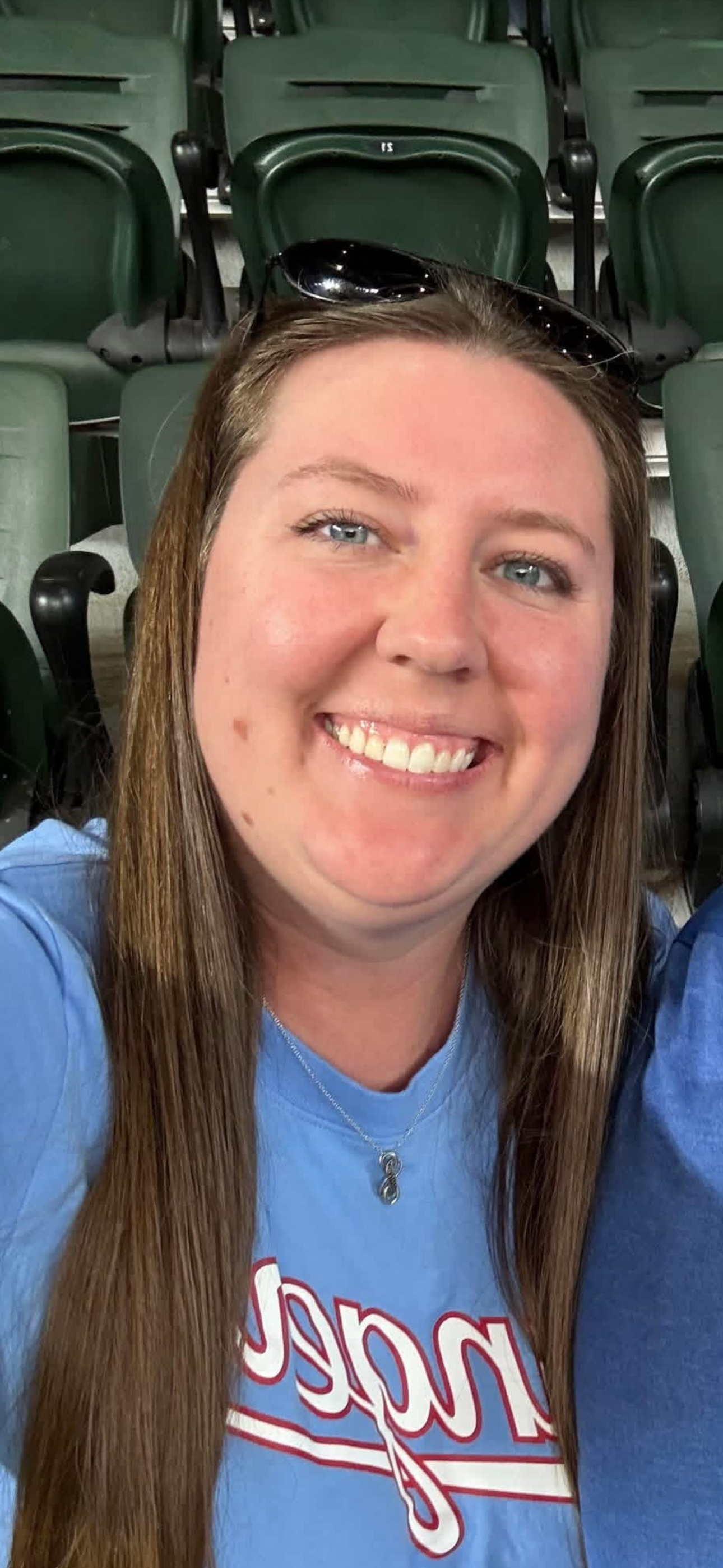 Close-up of a smiling woman with long brown hair, blue eyes, and sunglasses on her head, sitting in an indoor stadium or arena with green seats in the background.