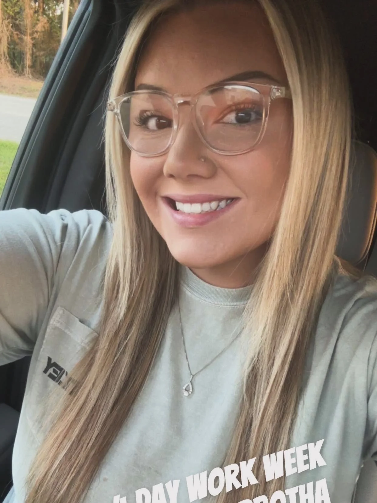 A smiling woman with long blonde hair wearing clear glasses, a gray T-shirt, and a silver necklace, sitting inside a vehicle with a view of trees outside.