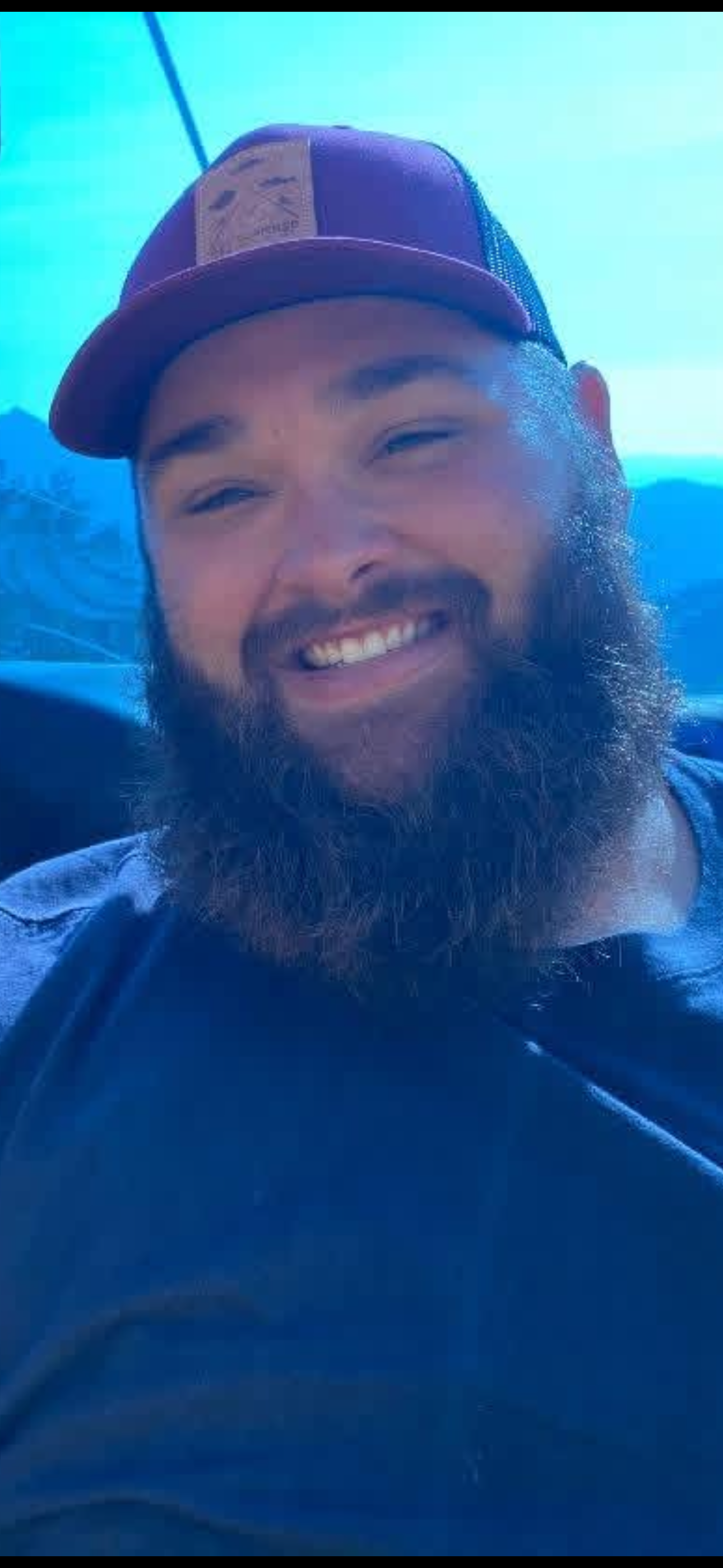 Close-up of a smiling man with a beard wearing a cap and a dark shirt, with a blue background.