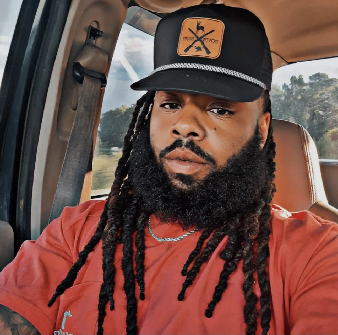 A man with a thick beard and long dreadlocks wearing a red shirt and black cap with a logo, sitting in a vehicle with a scenic landscape visible outside the window.